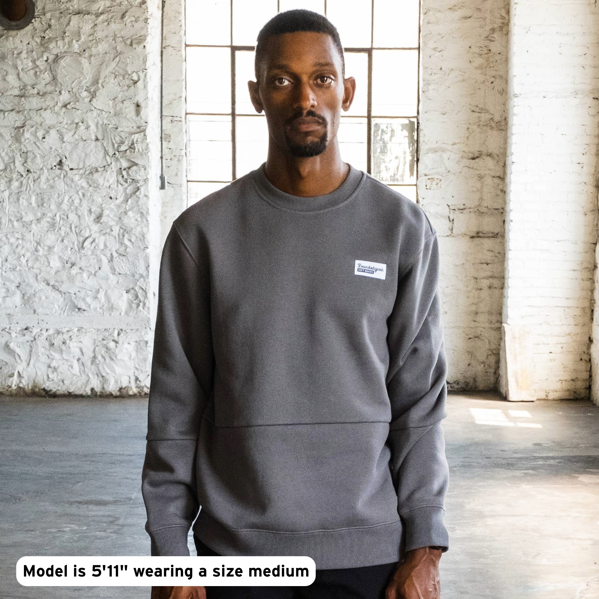 A man stands indoors by a large window, wearing the beardedgoat Men's Void Crew heavyweight cotton fleece sweatshirt in gray and black pants. Text: Model is 5'11" wearing a size medium. The room features white brick walls and natural light.