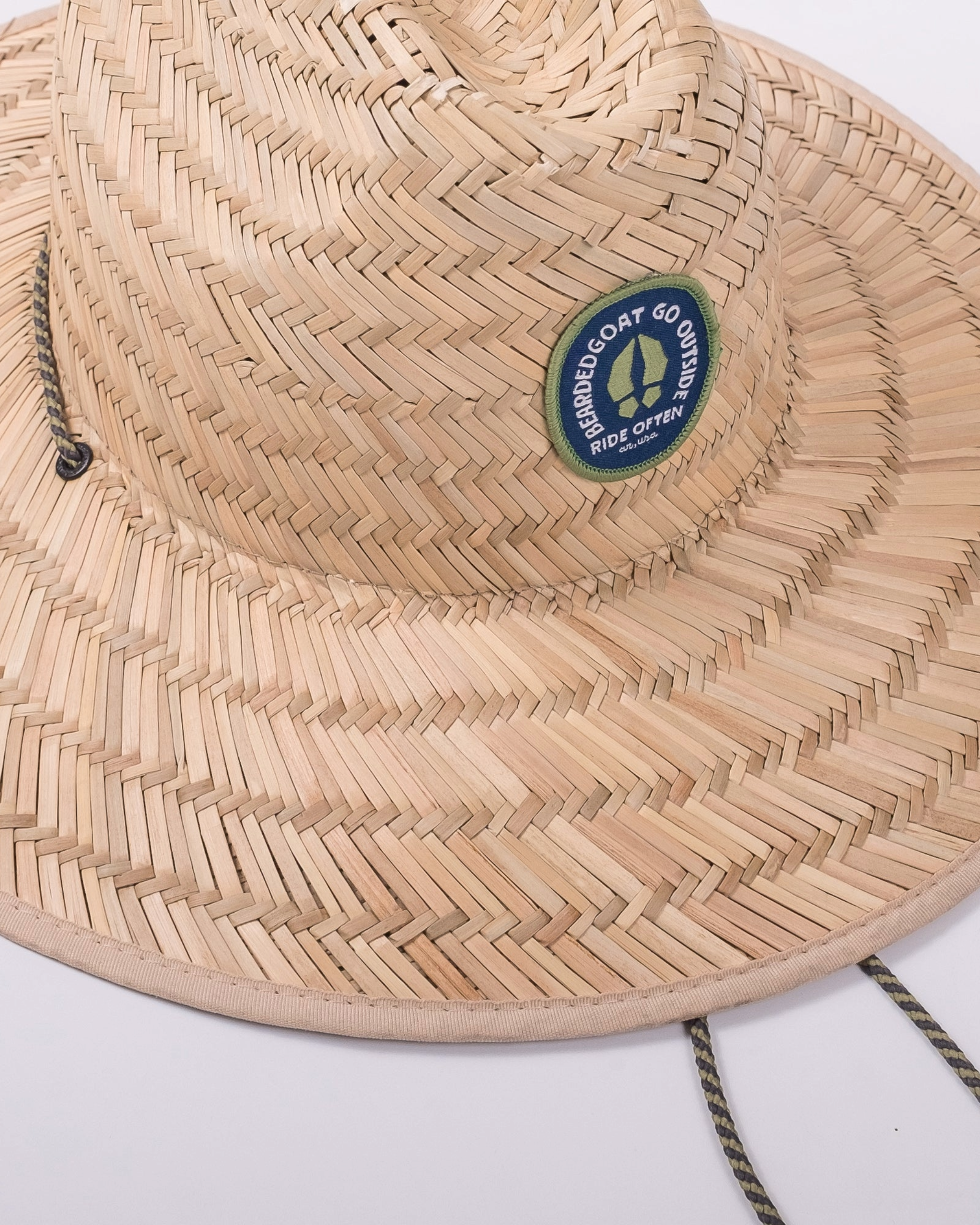 The beardedgoat Sun Hat is a woven straw wide-brim hat offering UV protection and a secure chin cord. It features a round patch with a goat silhouette and the phrase ROAMING GOAT GO OUTSIDE RIDE OFTEN.
