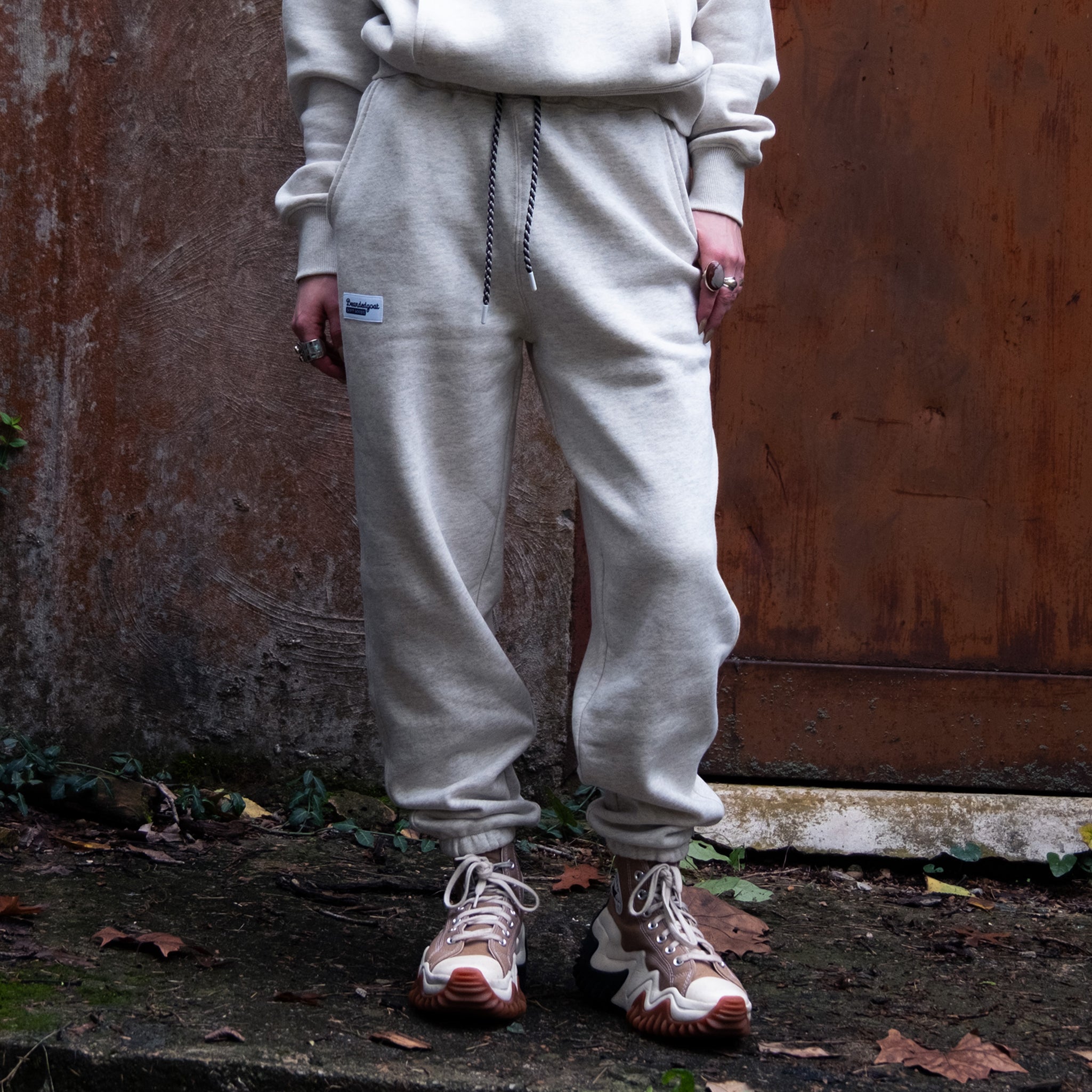 Wearing the NEW Overnight™ Classic Jogger by BEARDEDGOAT, paired with a matching sweatshirt and beige sneakers, a person stands on a concrete slab near a weathered brown door amid fallen leaves and greenery. Their face is not visible.