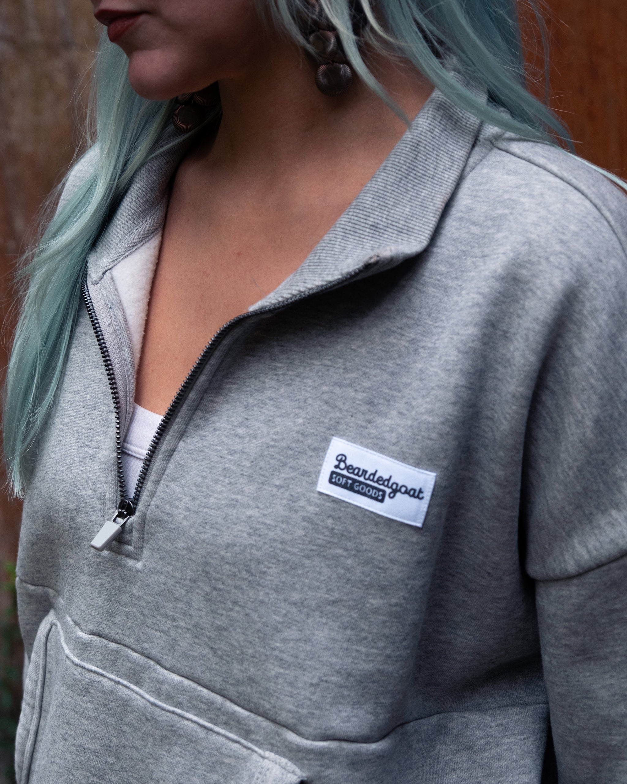 A person with long, light blue hair wears the BEARDEDGOAT NEW — Overnight™ Half Zip in heather grey. Part of their face is visible, and a blurred wooden outdoor background is seen behind them.