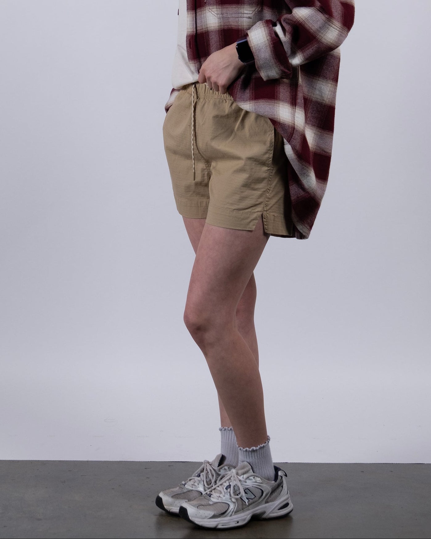 A woman with long dark hair, in profile against a plain background, wears BEARDEDGOAT Women's Canopy Shorts with stretch cotton ripstop, a red and white plaid shirt, gray socks, and white sneakers, looking slightly forward.
