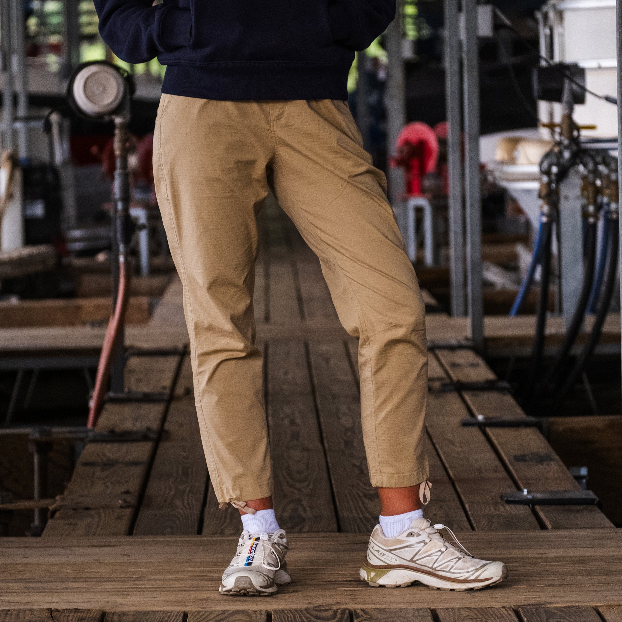 A person wearing BEARDEDGOAT Women's Canopy™ Pant with white socks and sneakers stands on a wooden walkway in an industrial setting, their upper body mostly out of frame.