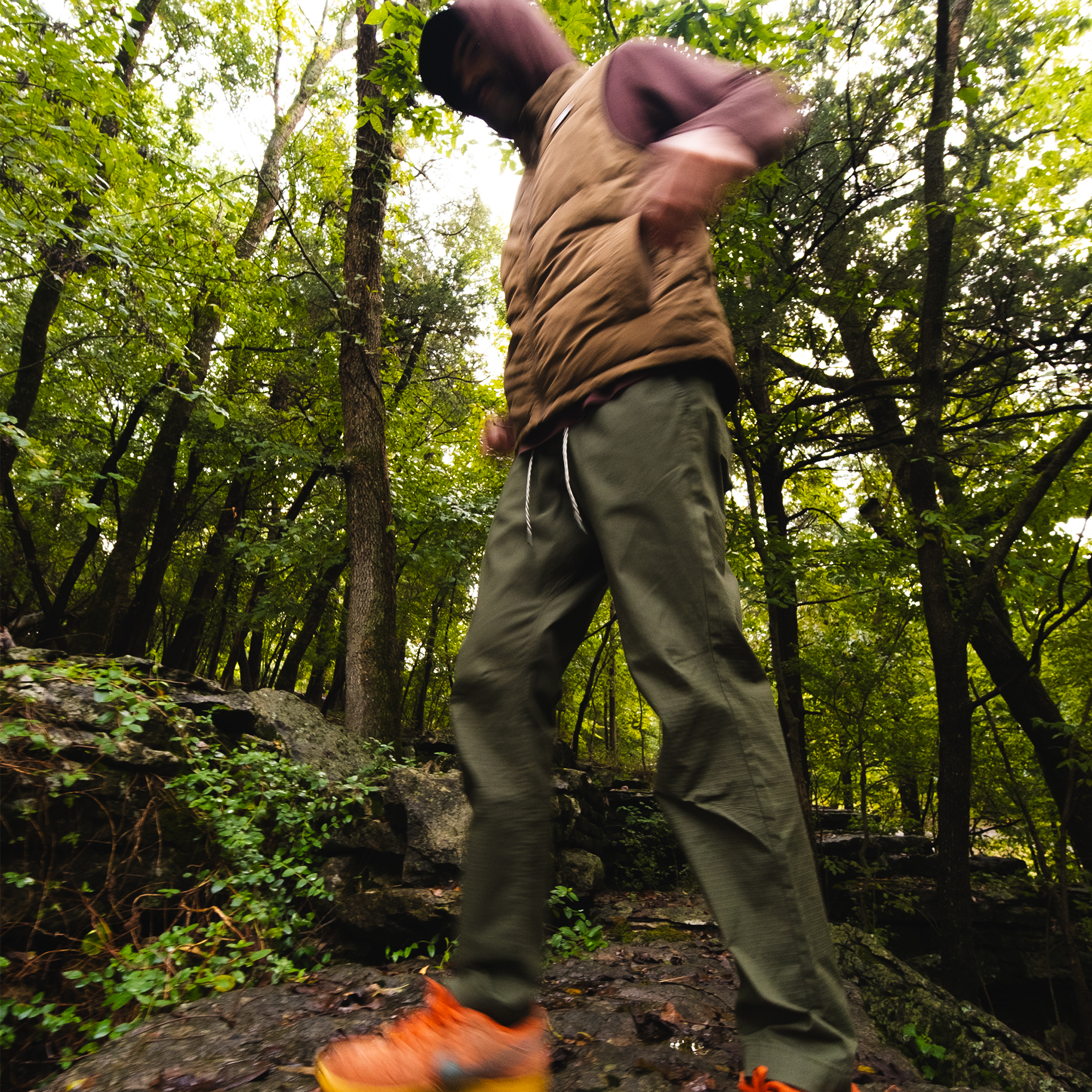 Wearing the BEARDEDGOAT Men's Canopy Pant, a person walks energetically along a forest trail lined with lush green trees. The image is slightly blurred, highlighting their motion through nature.