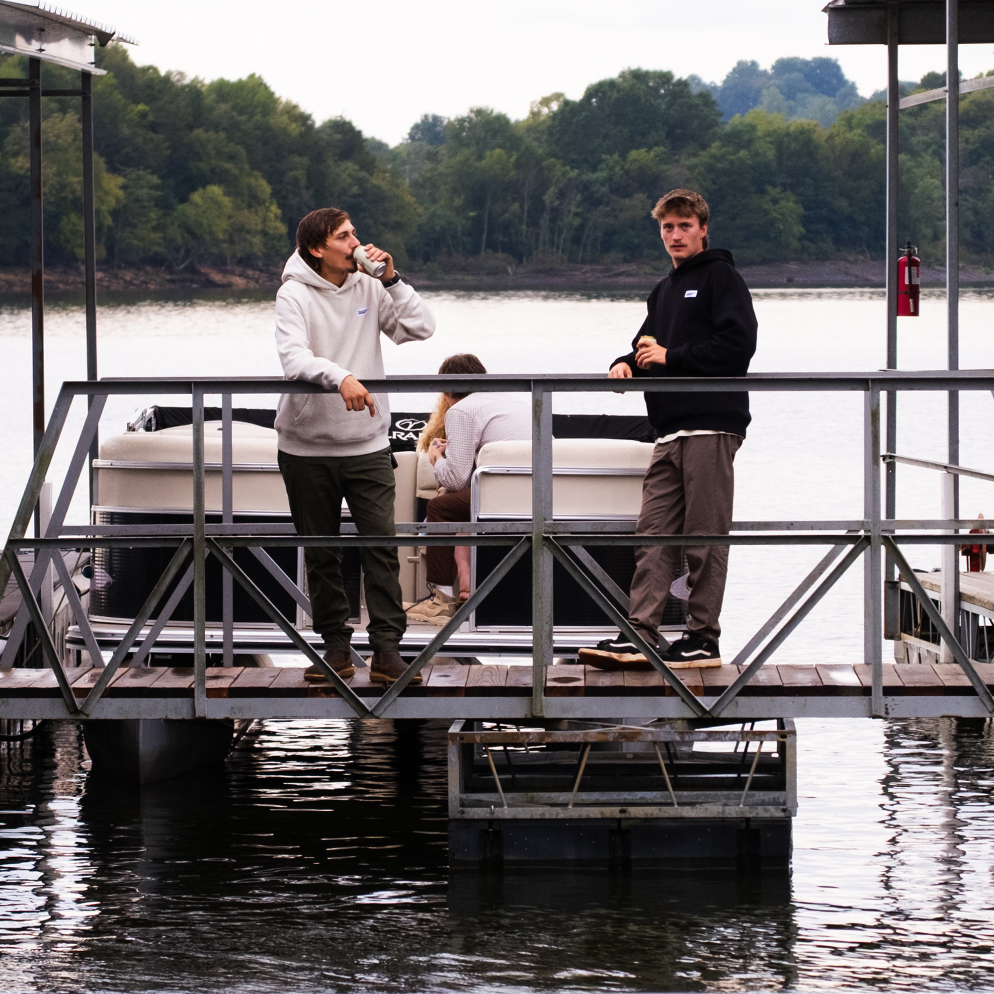 Two men stand on a dock by a pontoon boat; one is on the phone, the other faces the camera. Both look adventure-ready in BEARDEDGOAT Men's Canopy Pants. Another person sits on the boat. Trees and water form the background.