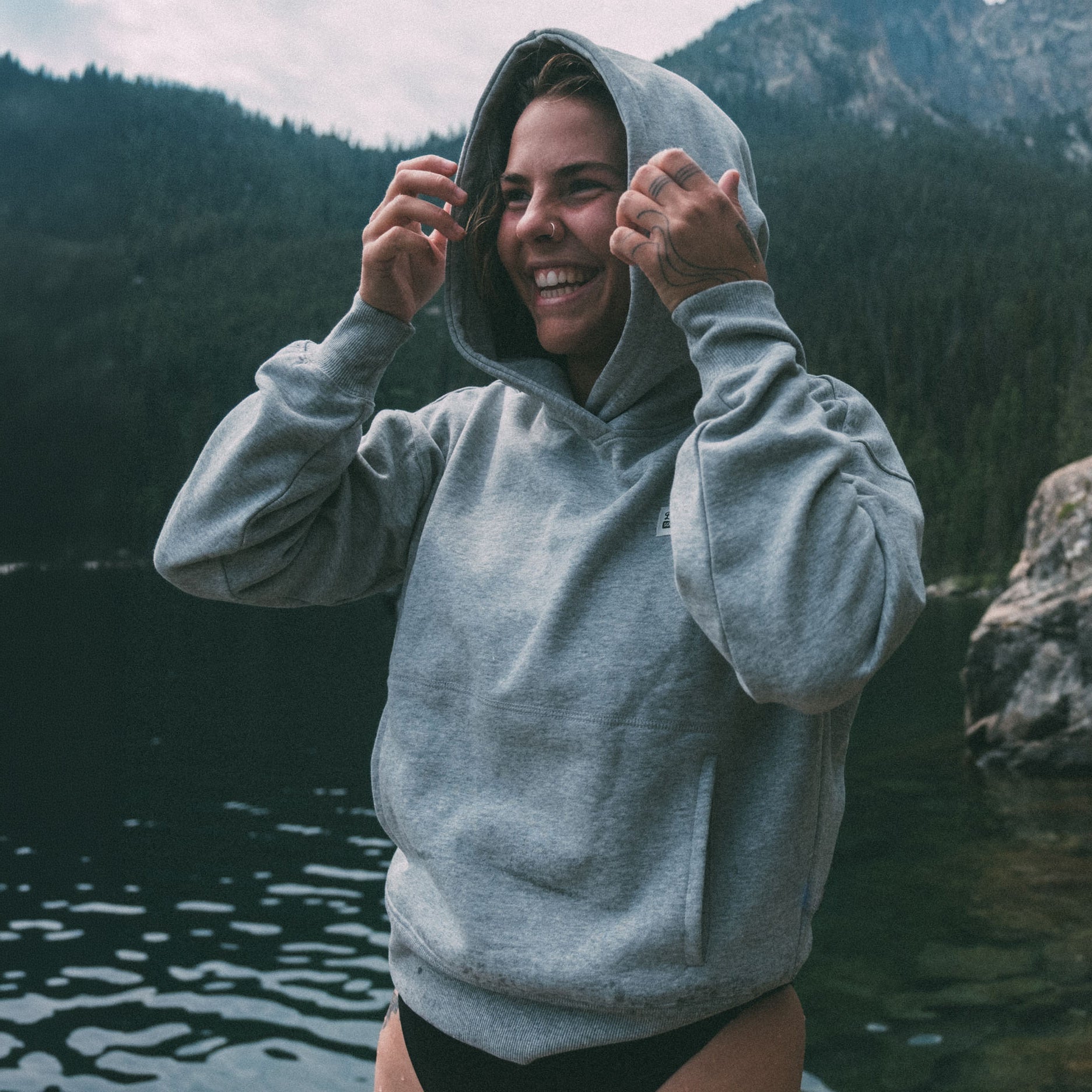 A smiling woman wears the beardedgoat Women's Overnight™ Hoodie with a relaxed drape by a lake, pulling up the hood. Forested mountains and rocky cliffs are in the background.