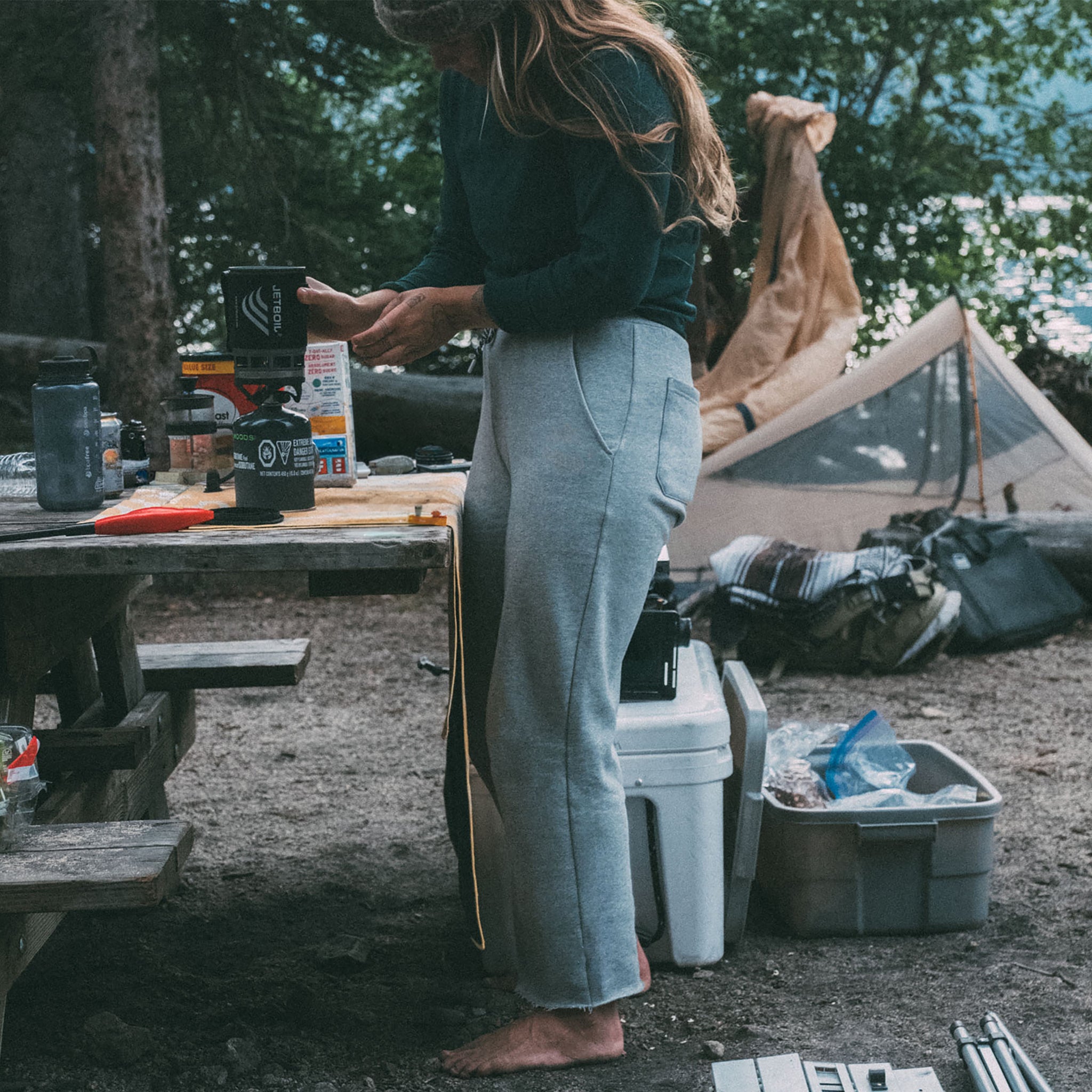 At a campsite among trees, a person with long hair stands barefoot at a wooden picnic table preparing food, wearing beardedgoat Women’s Overnight Sweatpant. Camping gear, a cooler, and a tent are visible in the background.
