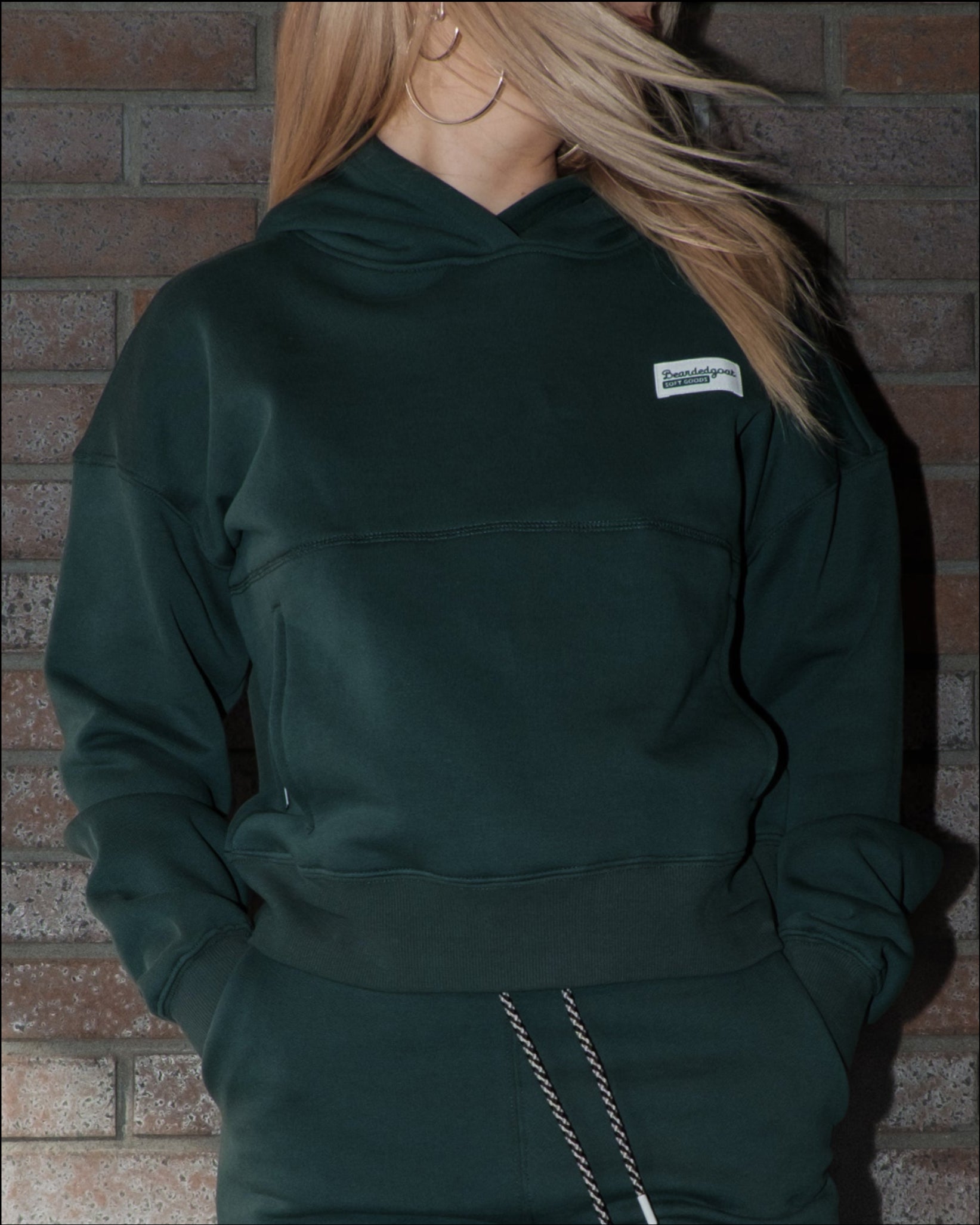 A person with long blonde hair stands against a brick wall in a dark green beardedgoat Women's Overnight™ Hoodie and matching sweatpants. Their face is partly turned away, partially hidden by their hair.