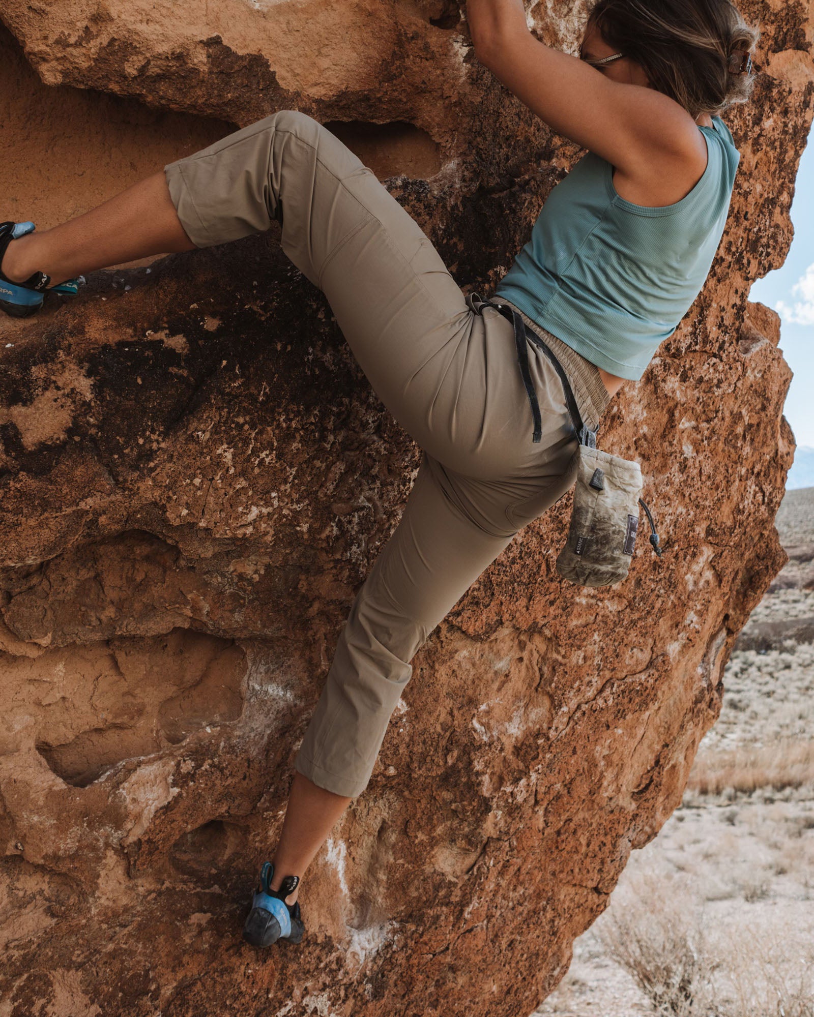 A woman climbs a rugged outdoor rock in BEARDEDGOAT’s NEW — Summit™ Pant and a blue tank top, with a chalk bag at her waist and an arid landscape behind her.