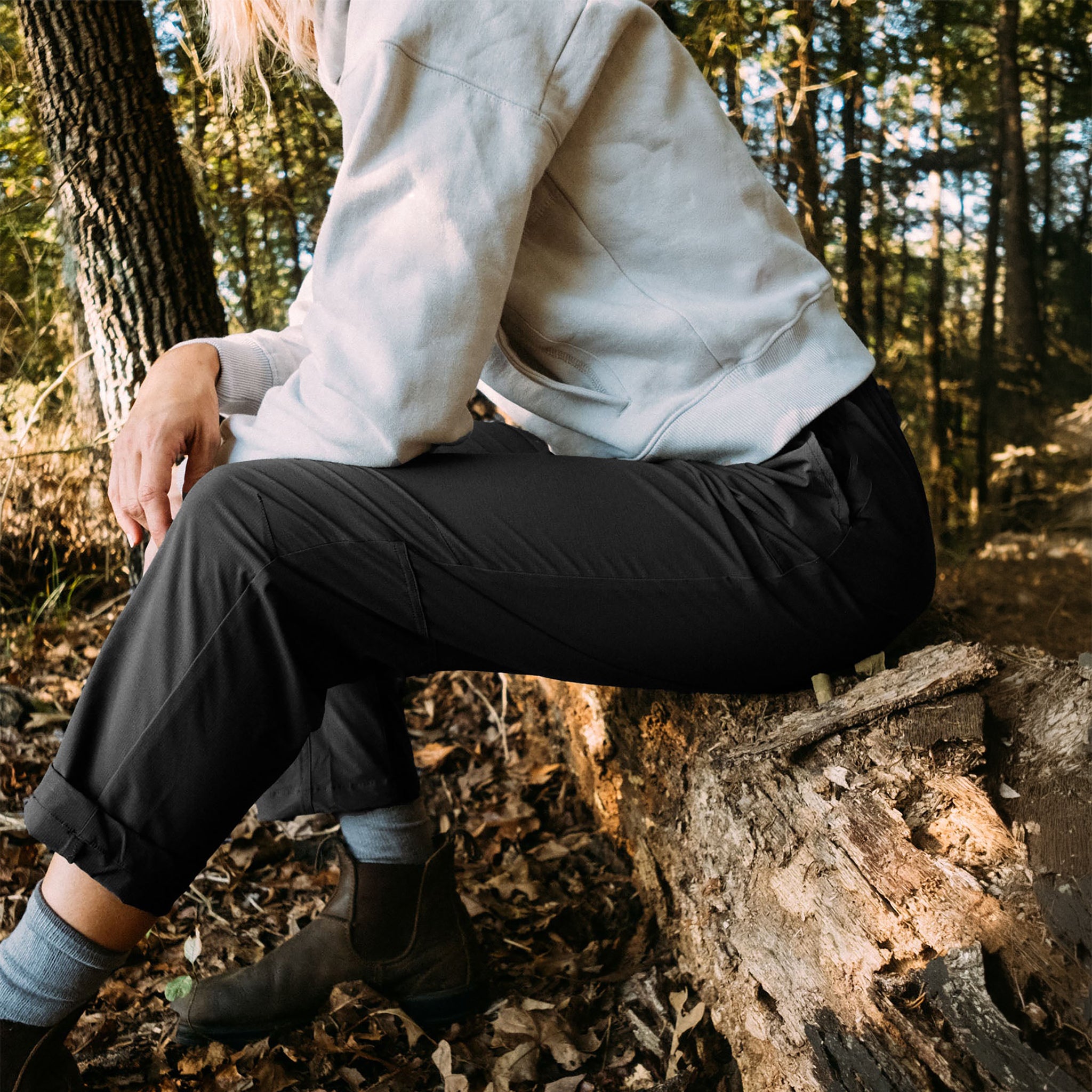 Wearing the BEARDEDGOAT NEW — Summit™ Pant and a white sweatshirt, a person sits on a fallen log in a sunlit forest, surrounded by trees and leaves.