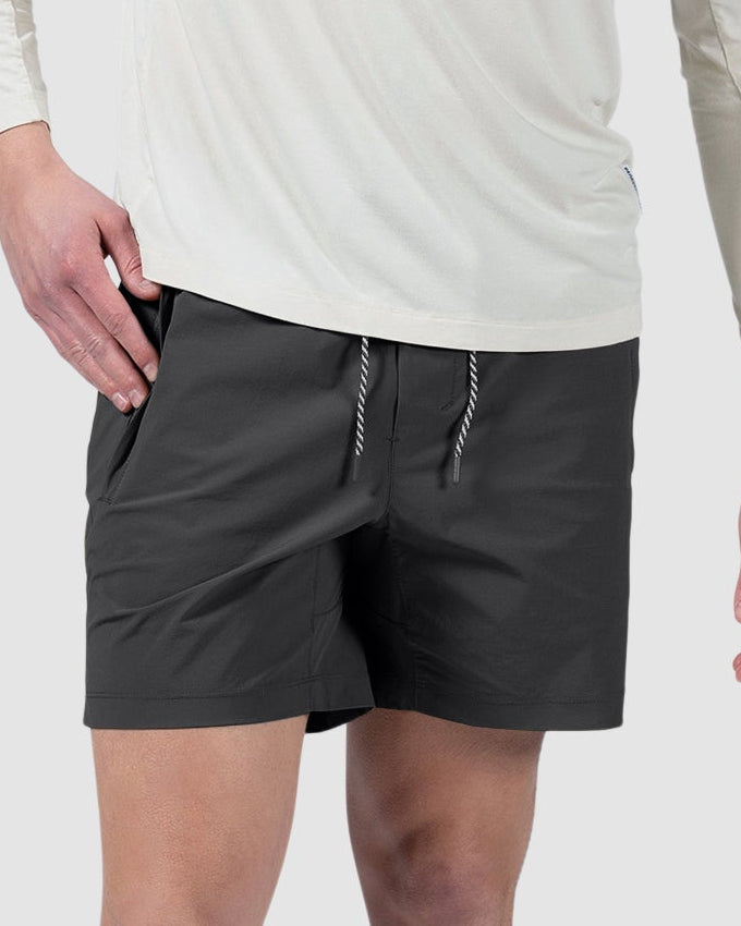 A man with short hair stands against a plain light gray background wearing BEARDEDGOAT Mens Rambler Short, a long-sleeve white shirt, white socks, and sneakers. The shorts feature 4-way stretch for comfort. He looks slightly left with one hand in his pocket.