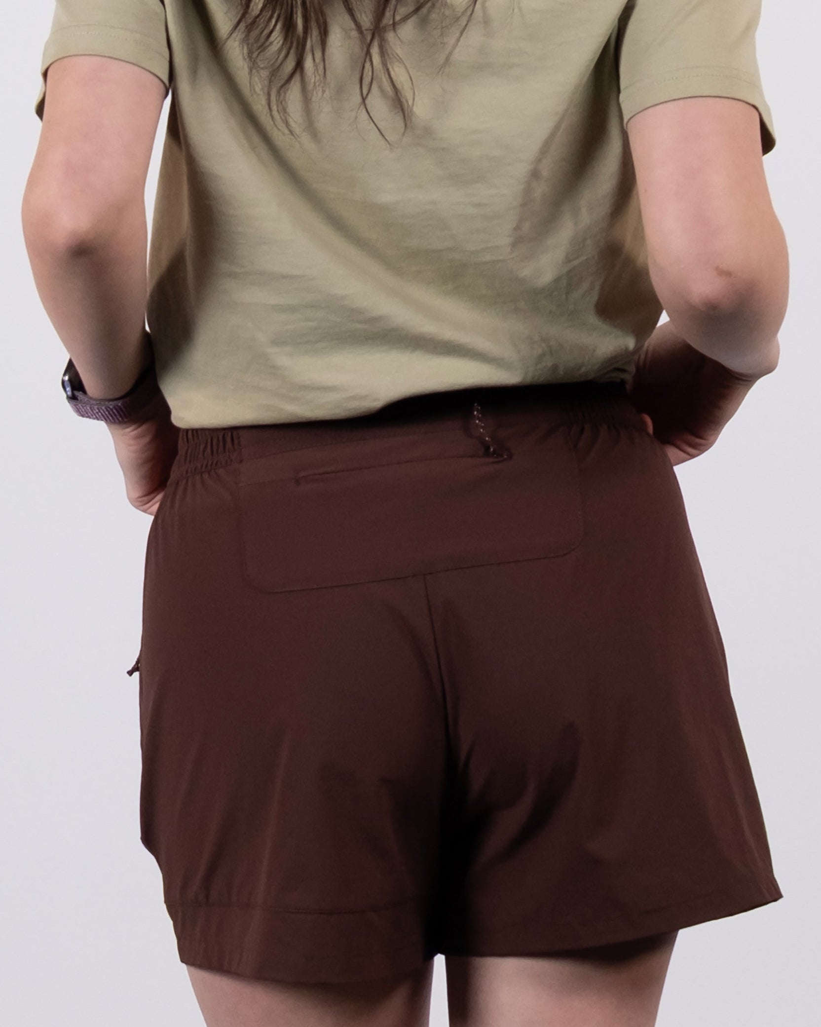 A person with long hair, wearing a short-sleeved beige shirt and BEARDEDGOAT Women's Alpina Shorts in brown, is shown from the back against a plain light background.