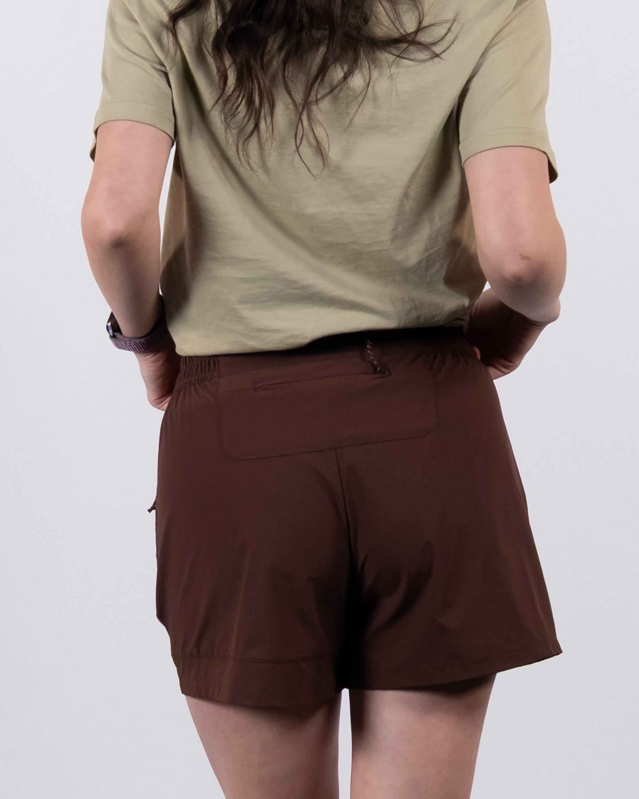 A person with long hair, wearing a short-sleeved beige shirt and BEARDEDGOAT Women's Alpina Shorts in brown, is shown from the back against a plain light background.
