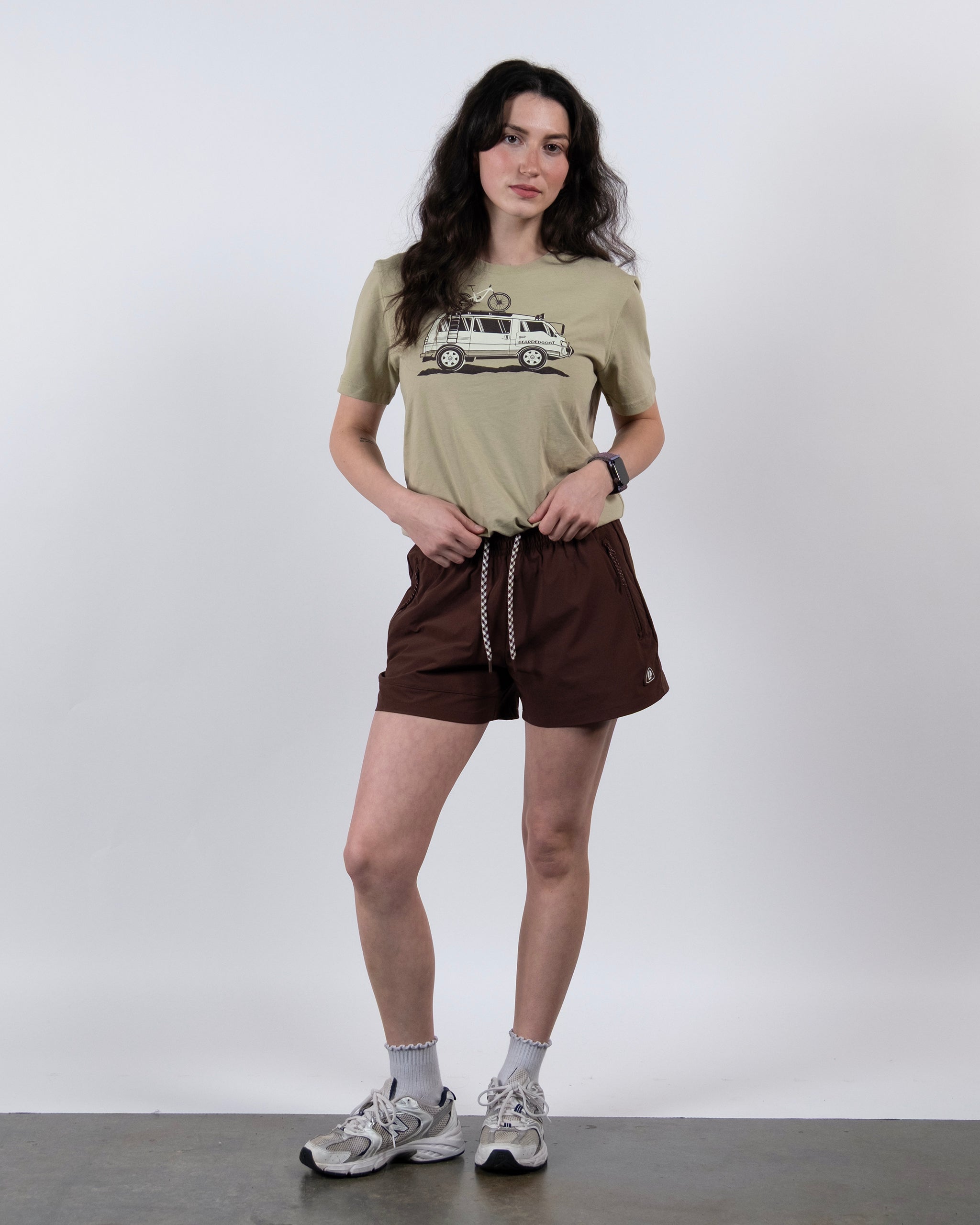 A woman with long wavy hair stands in a relaxed pose, wearing BEARDEDGOAT's Women's Alpina Short in brown ripstop, a beige car graphic tee, white socks, and gray sneakers against a plain background. One hand is in her pocket.