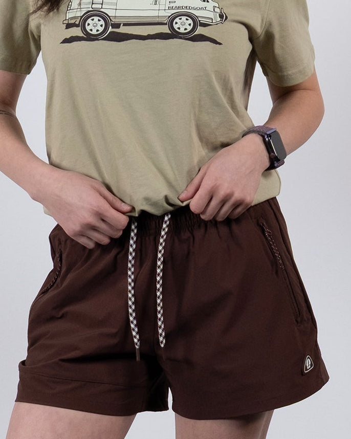 A woman with long wavy hair stands in a relaxed pose, wearing BEARDEDGOAT's Women's Alpina Short in brown ripstop, a beige car graphic tee, white socks, and gray sneakers against a plain background. One hand is in her pocket.