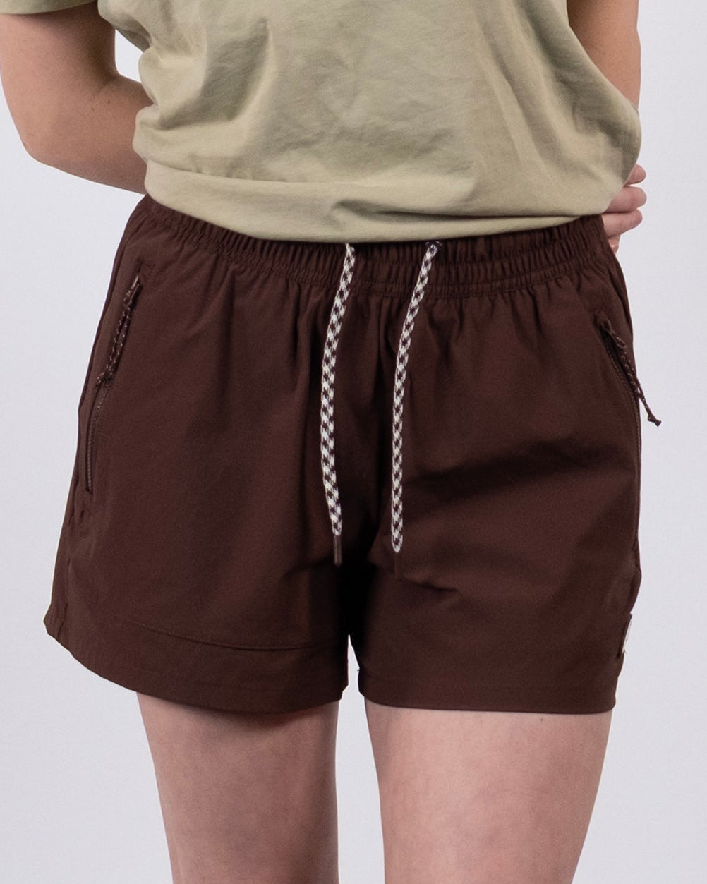 A person poses against a plain background, wearing the BEARDEDGOAT Women's Alpina Short in Chickory with a beige T-shirt, white socks, and sneakers. Their arms are crossed behind their back, and their head is out of frame.