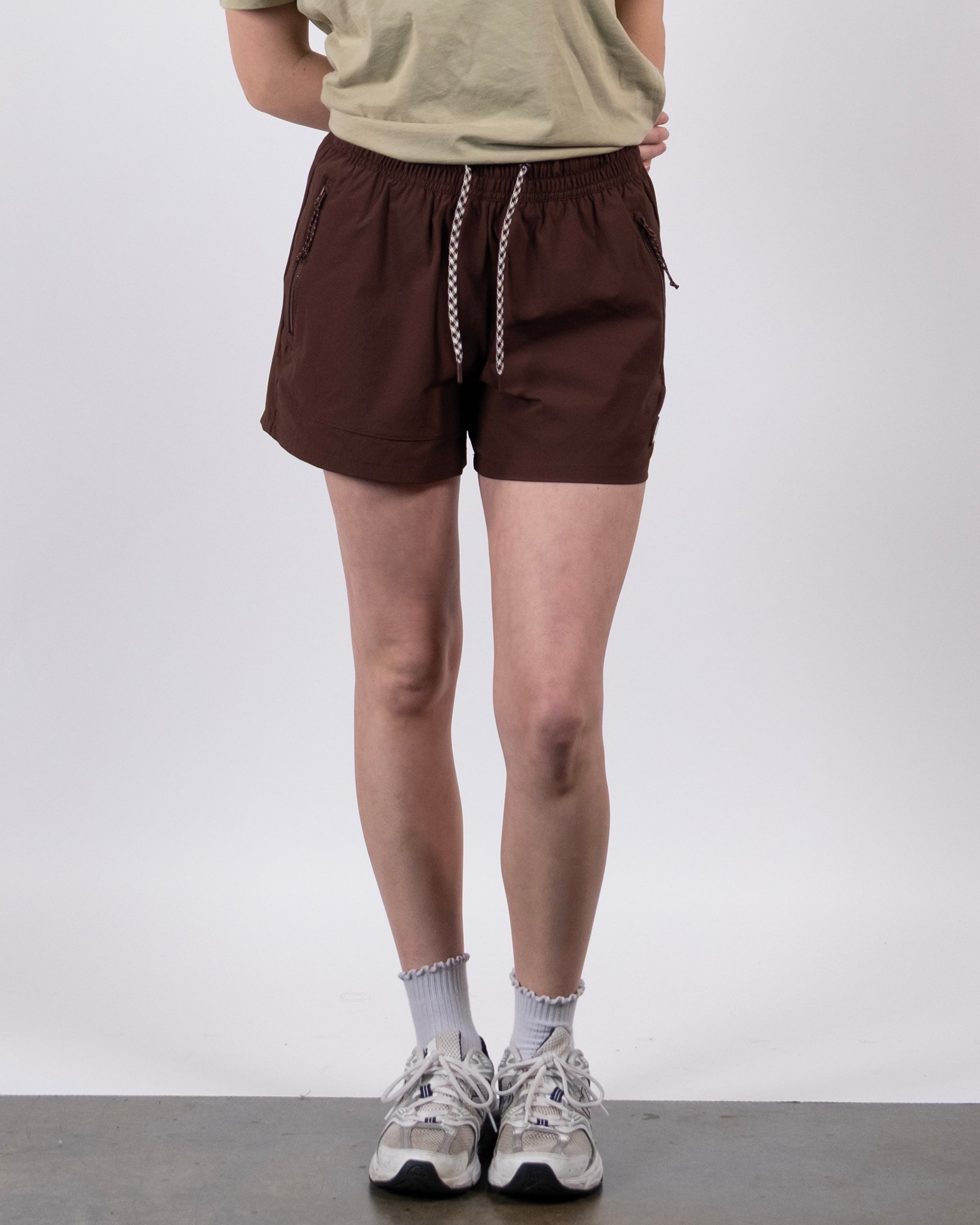 A person poses against a plain background, wearing the BEARDEDGOAT Women's Alpina Short in Chickory with a beige T-shirt, white socks, and sneakers. Their arms are crossed behind their back, and their head is out of frame.