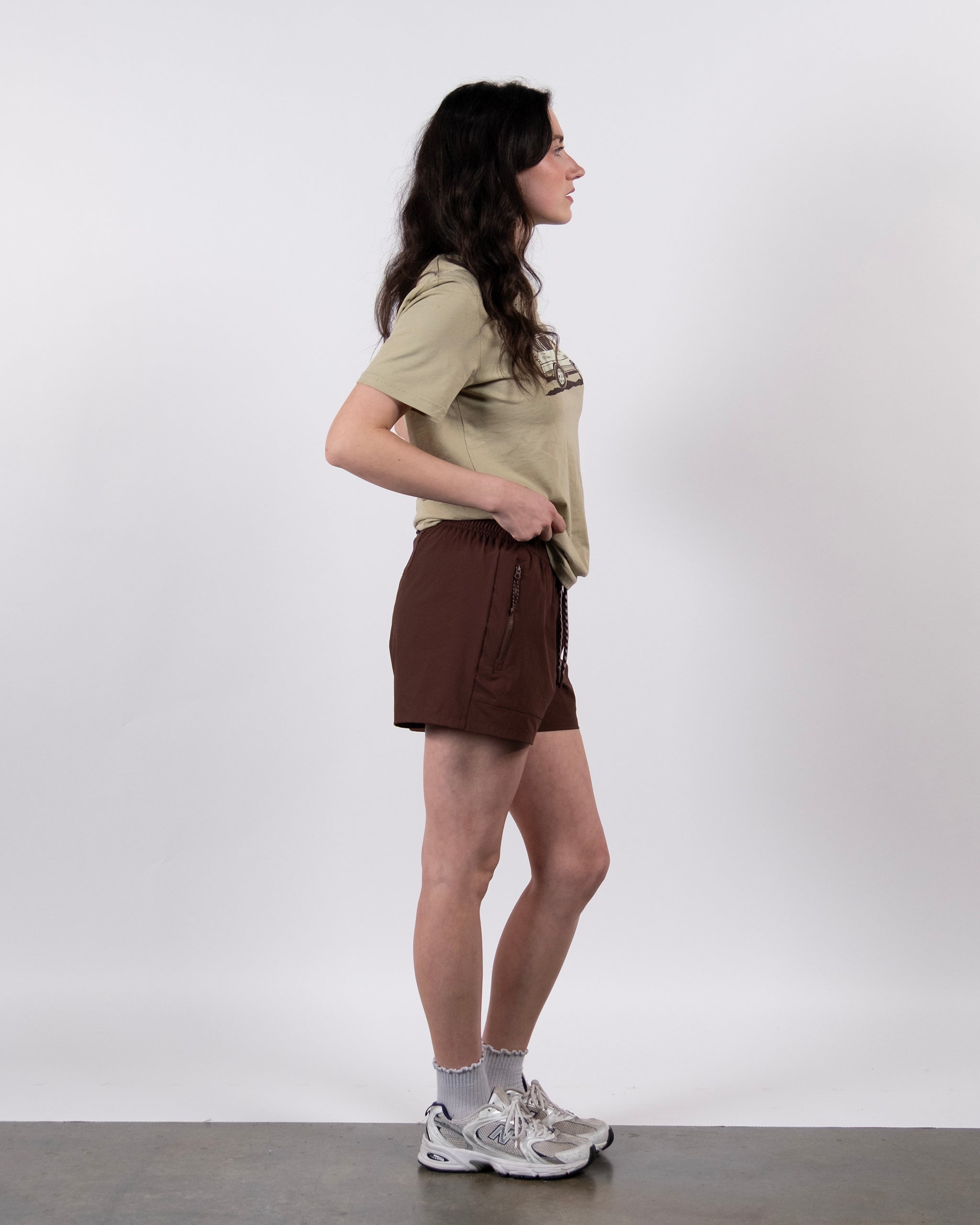 A woman stands in profile against a plain white background, wearing a beige t-shirt and BEARDEDGOAT Women's Alpina Short, which features durable ripstop fabric, paired with gray socks and white athletic shoes.