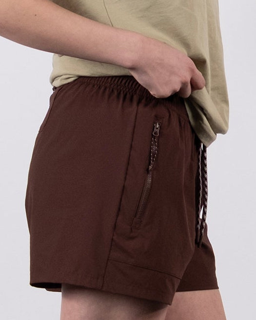 A woman stands in profile against a plain white background, wearing a beige t-shirt and BEARDEDGOAT Women's Alpina Short, which features durable ripstop fabric, paired with gray socks and white athletic shoes.