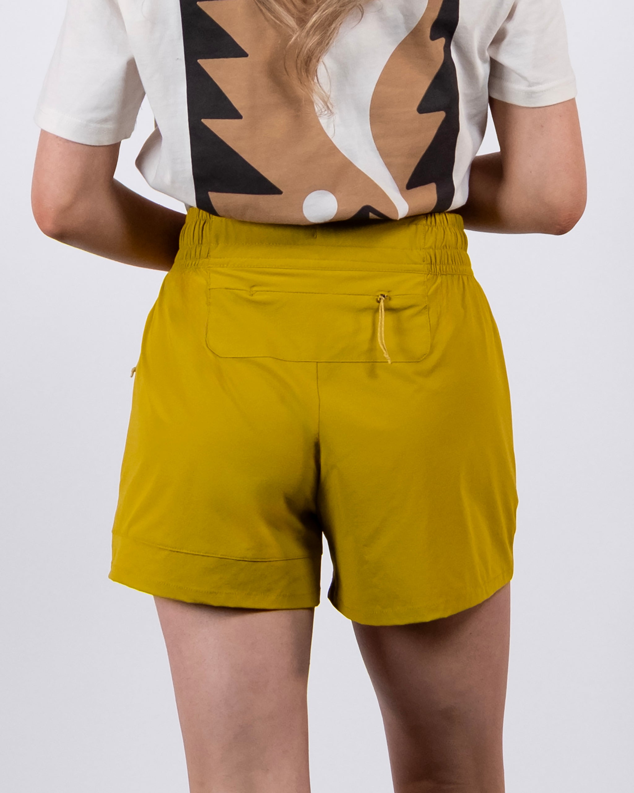 A person seen from the back wears BEARDEDGOAT Women's Alpina Short in mustard yellow ripstop with a back zip pocket and a white T-shirt featuring a bold abstract design.