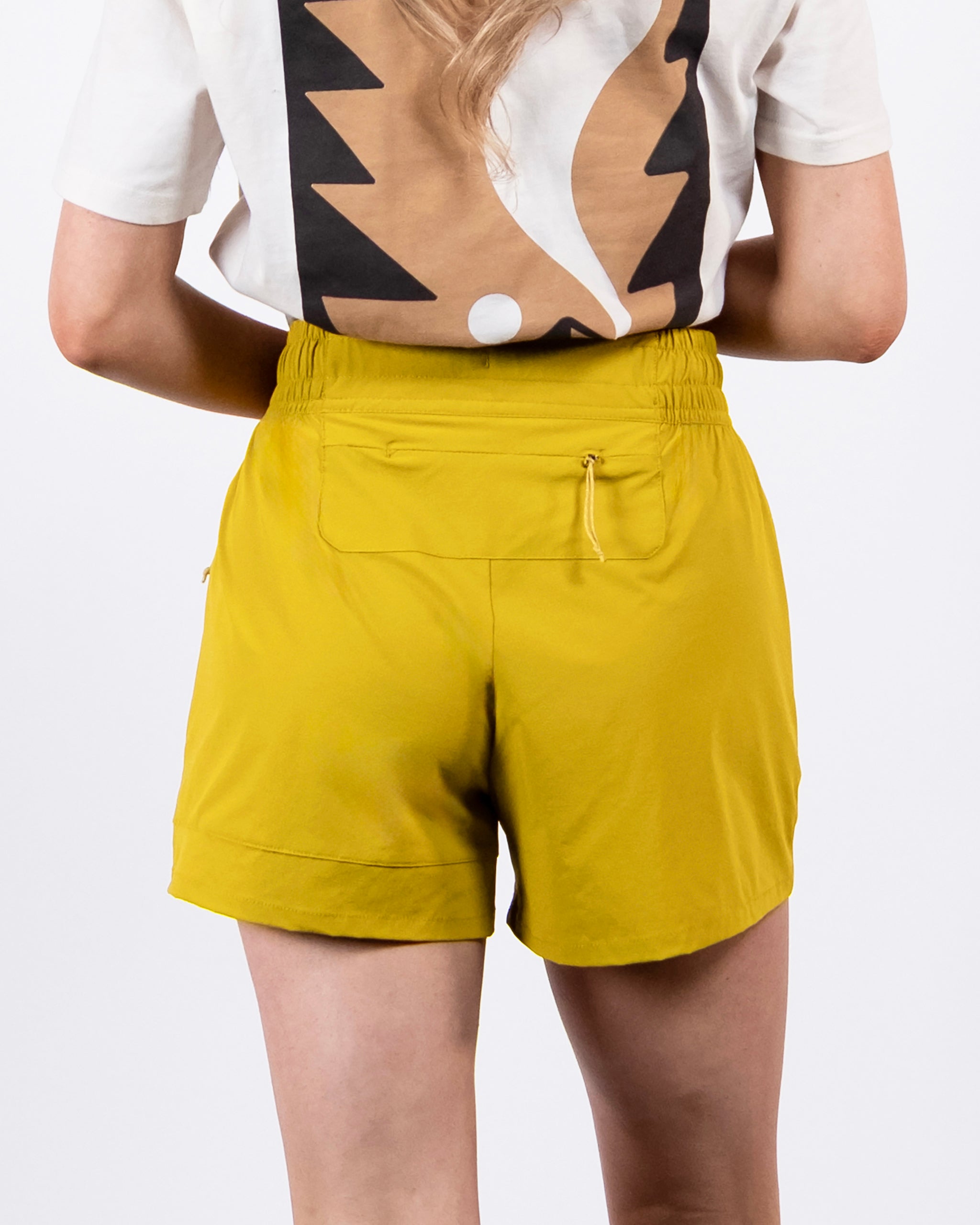 A person with long, wavy light brown hair stands facing away, wearing a white T-shirt with a bold abstract design and BEARDEDGOAT Women's Alpina Shorts—high-waisted adventure shorts in lightweight ripstop with a zippered back pocket.