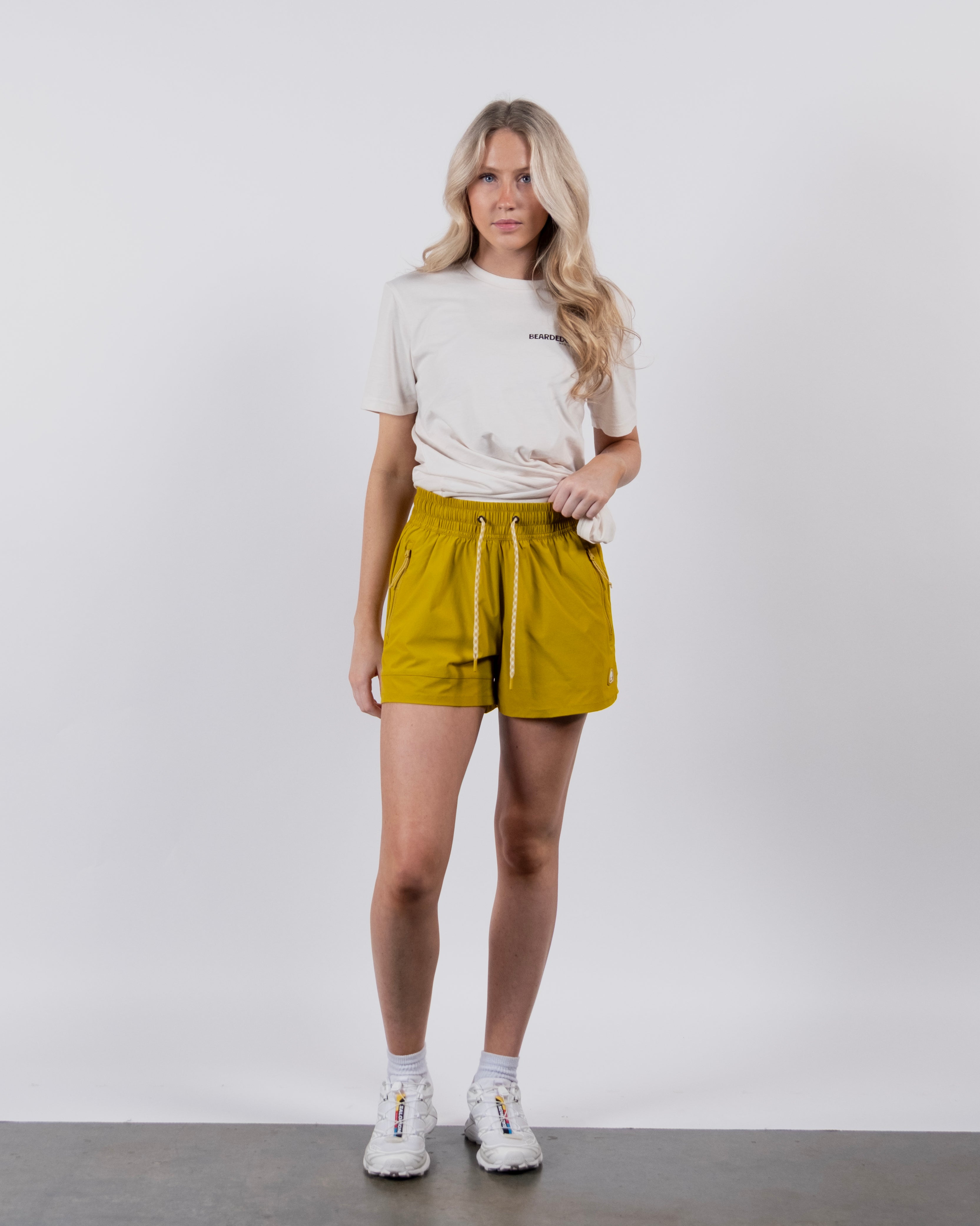 A woman with long blonde hair stands against a white background, wearing a light beige T-shirt and BEARDEDGOAT Women's Alpina Short in mustard yellow ripstop fabric with zippered pockets, paired with white sneakers. Her expression is neutral, left hand by her side.