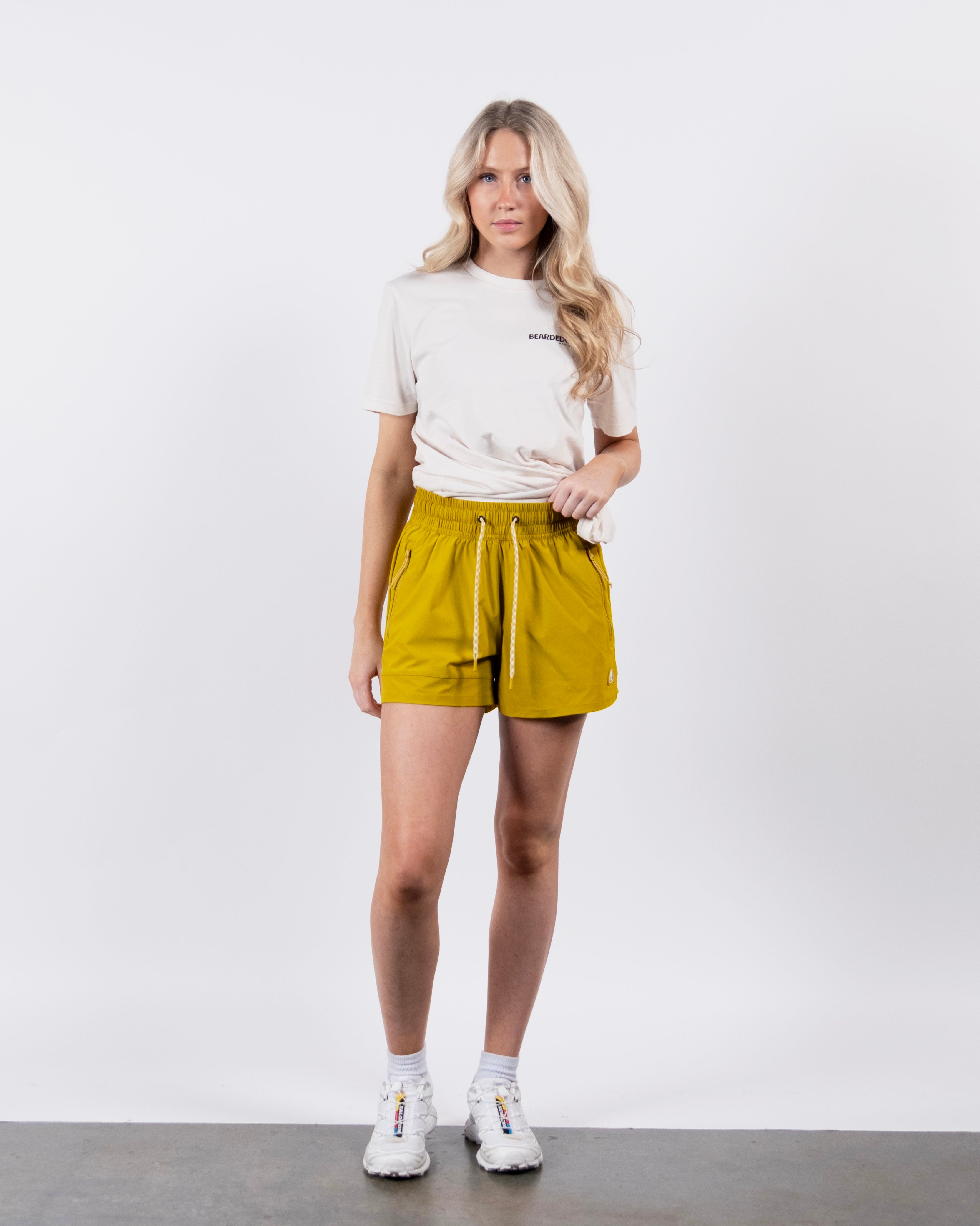 A woman with long blonde hair stands facing forward, wearing a white t-shirt and BEARDEDGOAT Women's Alpina Short in mustard yellow with zippered pockets, paired with white sneakers, against a plain white background.