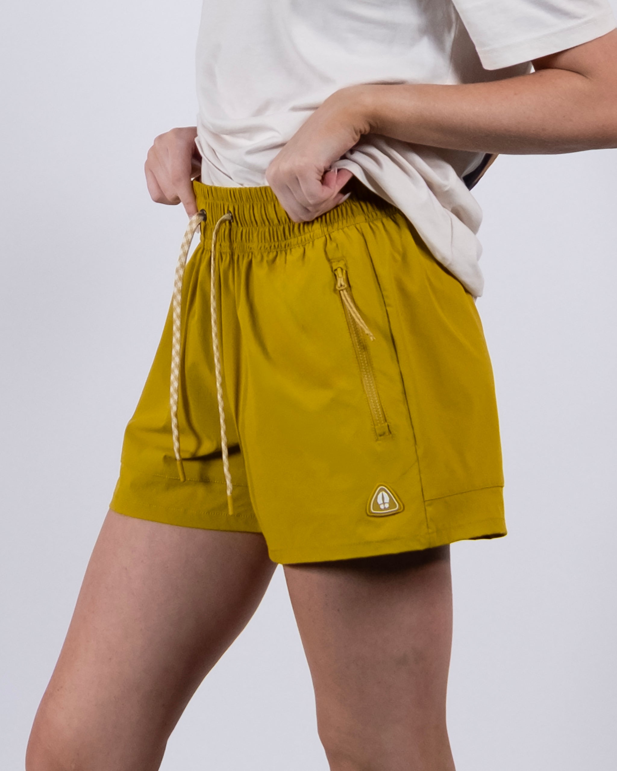 A person wearing a white t-shirt and BEARDEDGOAT Women's Alpina Short in mustard yellow with zippered pockets and a white drawstring, standing against a plain background.
