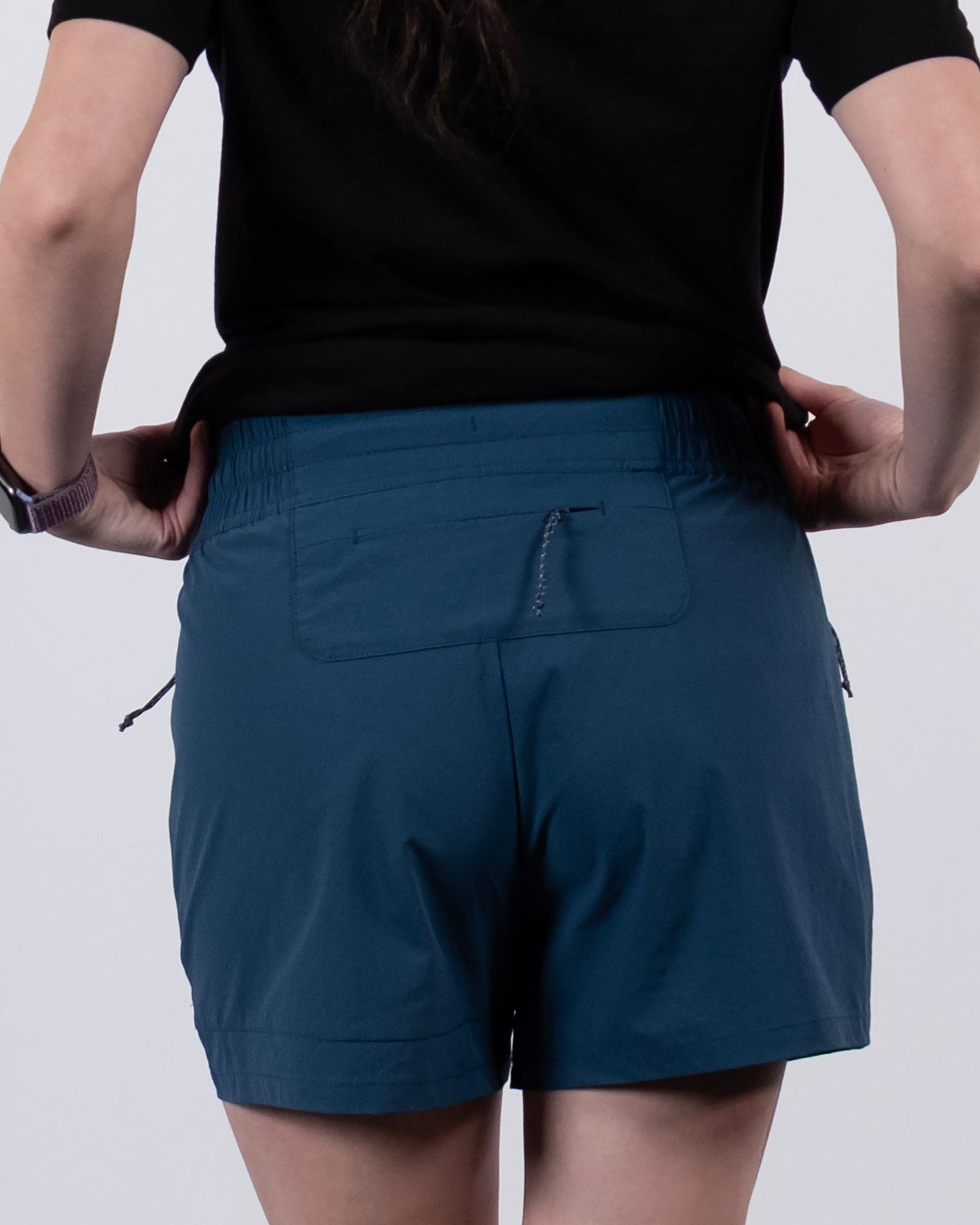 A person with long wavy hair, dressed in a black t-shirt and BEARDEDGOAT Women's Alpina Short, stands with their back to the camera against a plain white background.