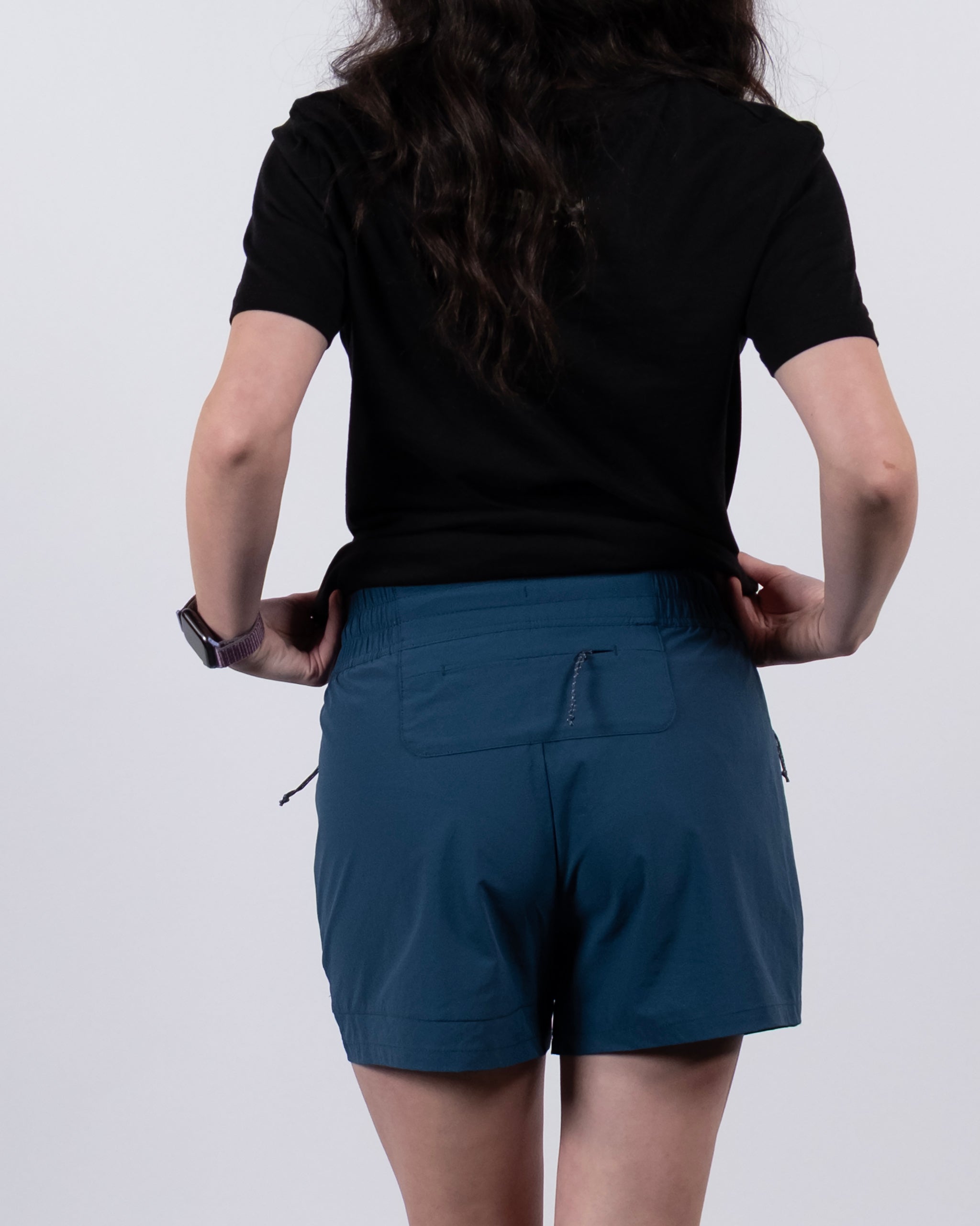 A person with long wavy hair, dressed in a black t-shirt and BEARDEDGOAT Women's Alpina Short, stands with their back to the camera against a plain white background.