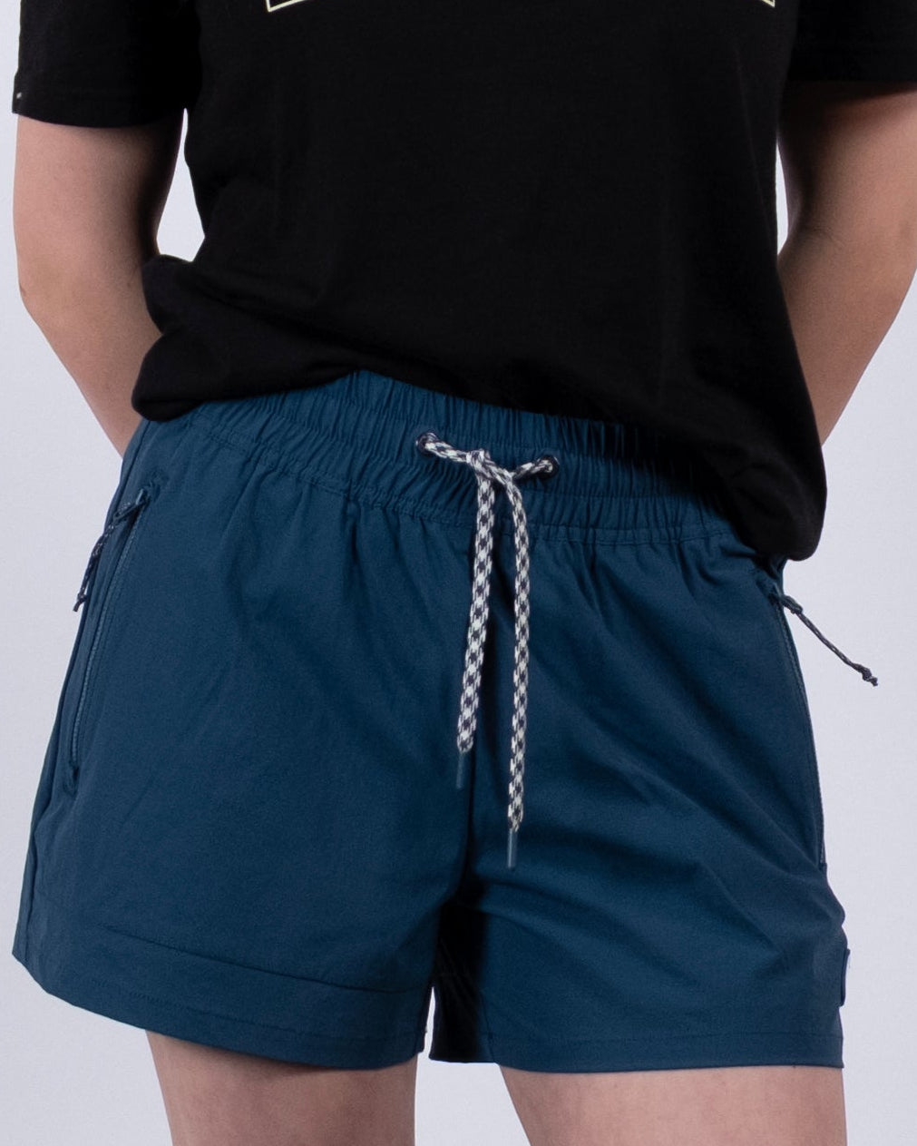 A person with long dark hair, glasses, a black graphic tee, BEARDEDGOAT Women's Alpina Short in blue ripstop, white socks, and sneakers stands against a plain white background with hands behind their back.