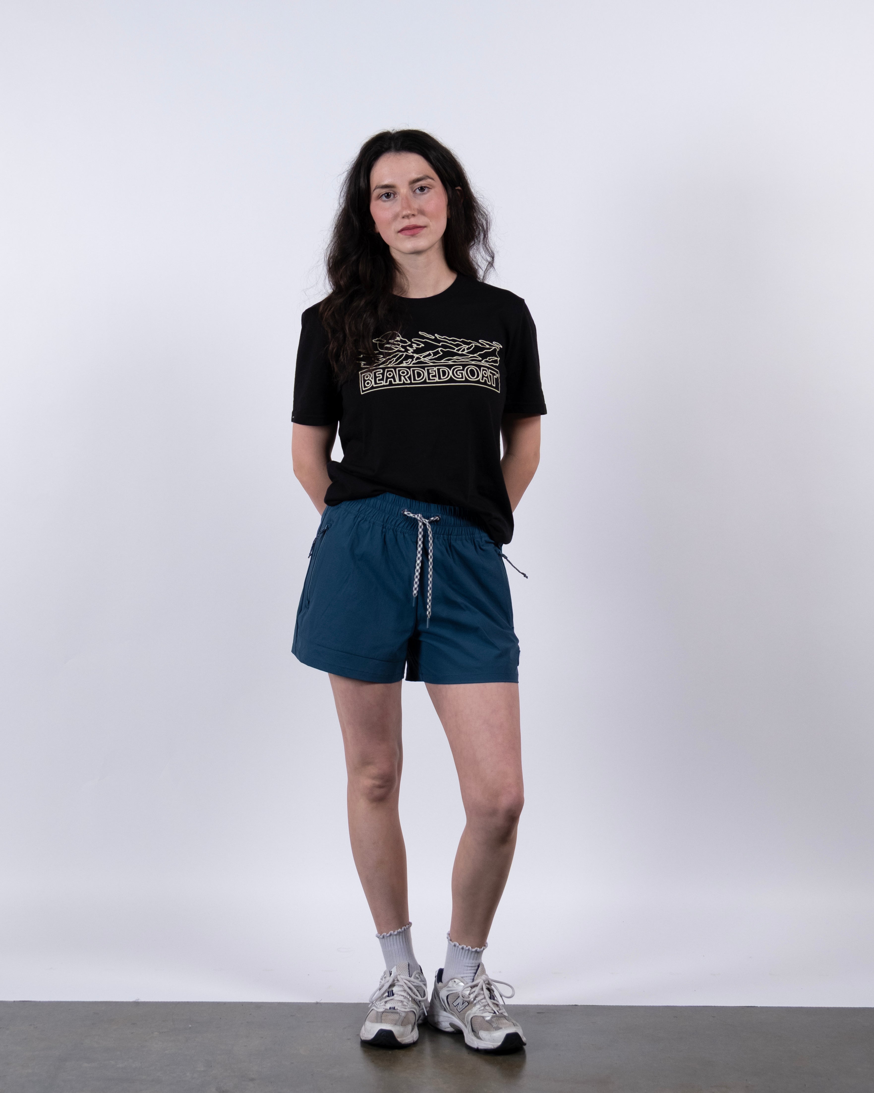 A person with long dark hair, glasses, a black graphic tee, BEARDEDGOAT Women's Alpina Short in blue ripstop, white socks, and sneakers stands against a plain white background with hands behind their back.