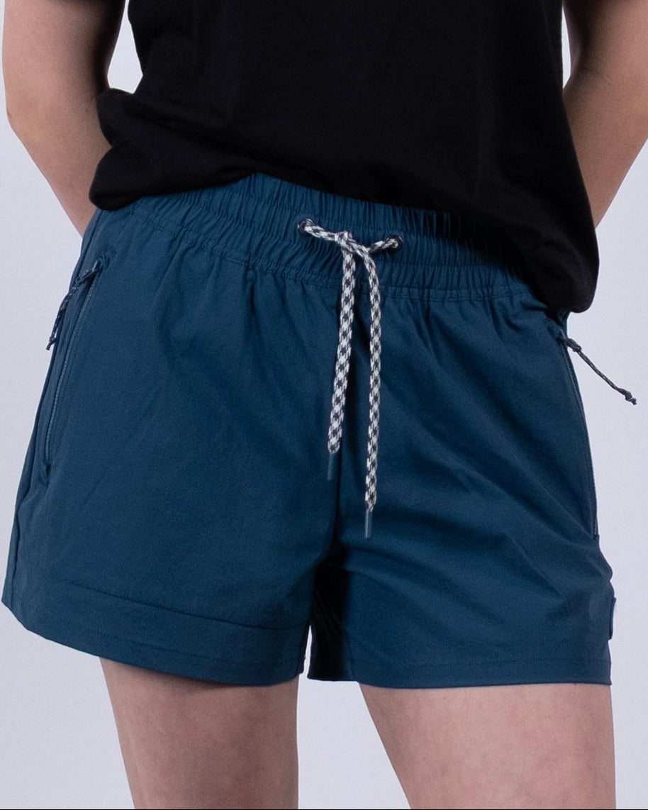 A person stands with hands behind their back, wearing a black T-shirt and BEARDEDGOAT Women's Alpina Short in teal with zippered pockets, along with white socks and gray sneakers against a plain light background.