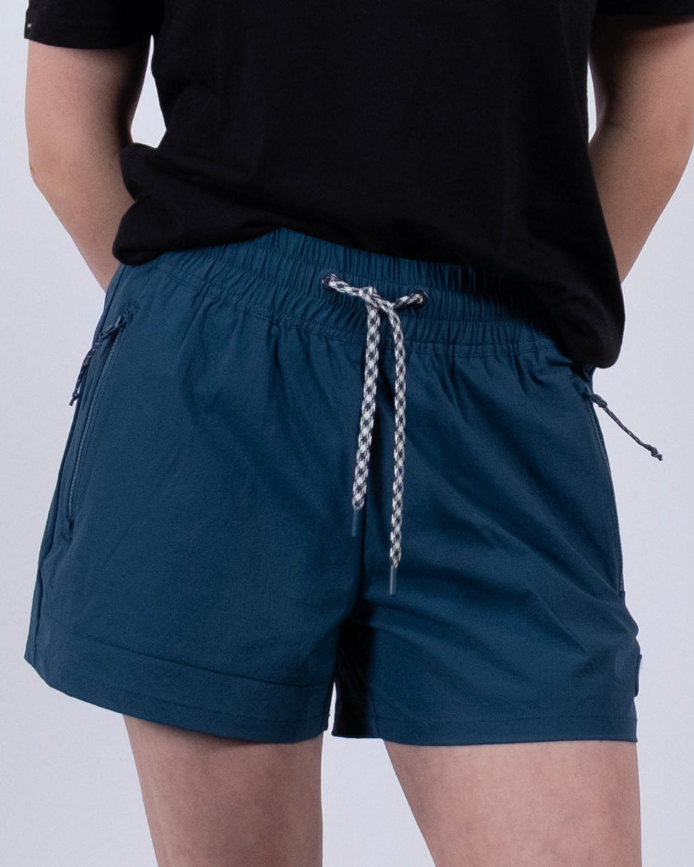 A person stands with hands behind their back, wearing a black T-shirt and BEARDEDGOAT Women's Alpina Short in teal with zippered pockets, along with white socks and gray sneakers against a plain light background.