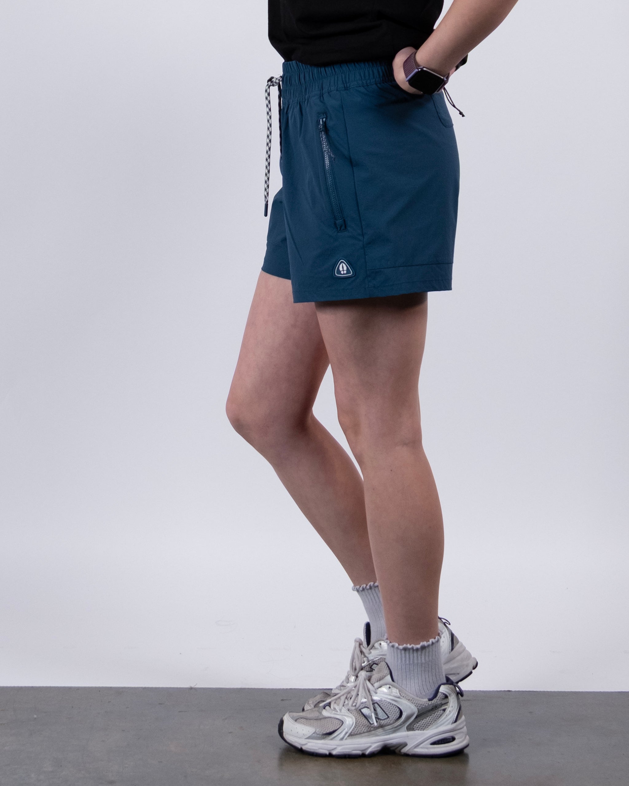 A person stands sideways in BEARDEDGOAT Women's Alpina Shorts (teal), a black top, white socks and sneakers, with a smartwatch. The background is plain and light-colored.