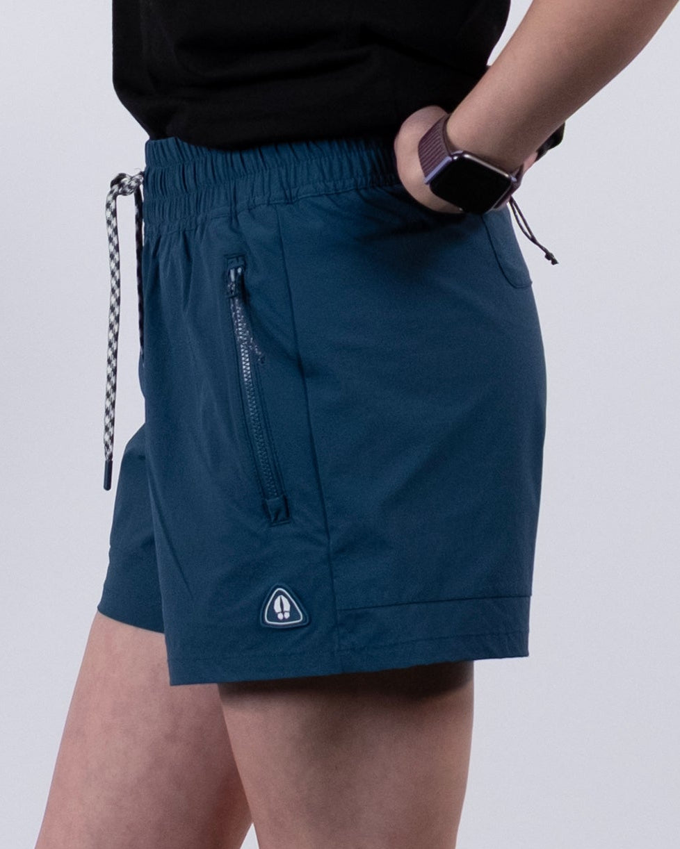 A person stands sideways in BEARDEDGOAT Women's Alpina Shorts (teal), a black top, white socks and sneakers, with a smartwatch. The background is plain and light-colored.