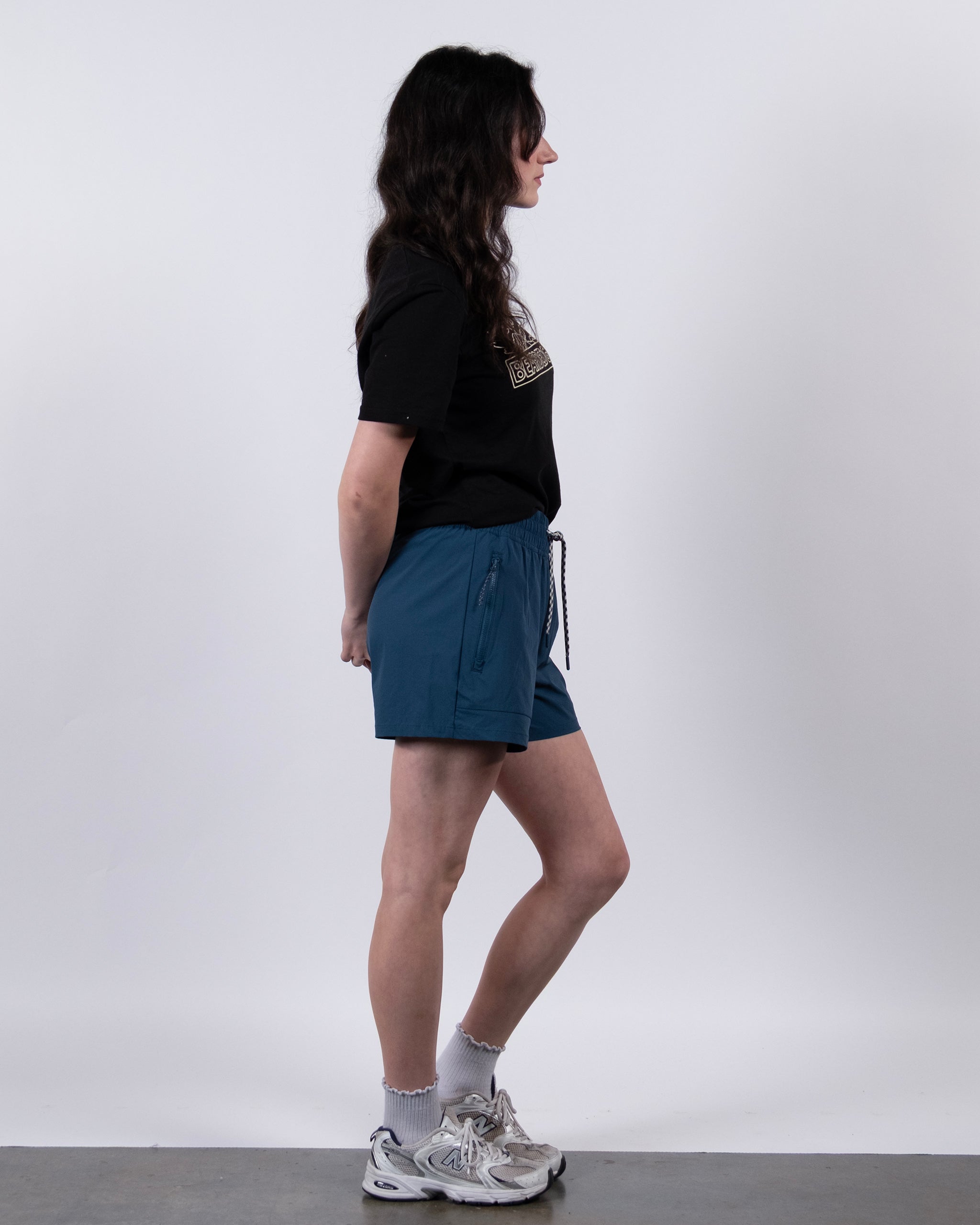 A person with long dark hair stands in profile against a plain background, wearing a black T-shirt, BEARDEDGOAT Women's Alpina ripstop teal shorts, gray socks, and white sneakers with hands behind their back.