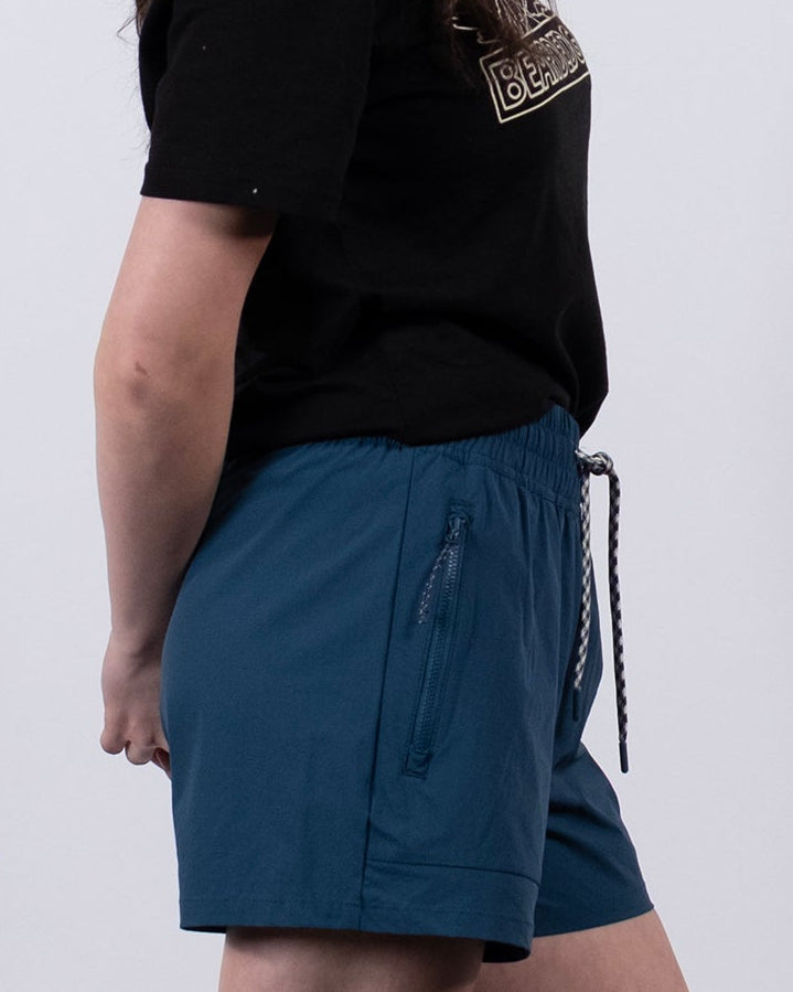 A person with long dark hair stands in profile against a plain background, wearing a black T-shirt, BEARDEDGOAT Women's Alpina ripstop teal shorts, gray socks, and white sneakers with hands behind their back.