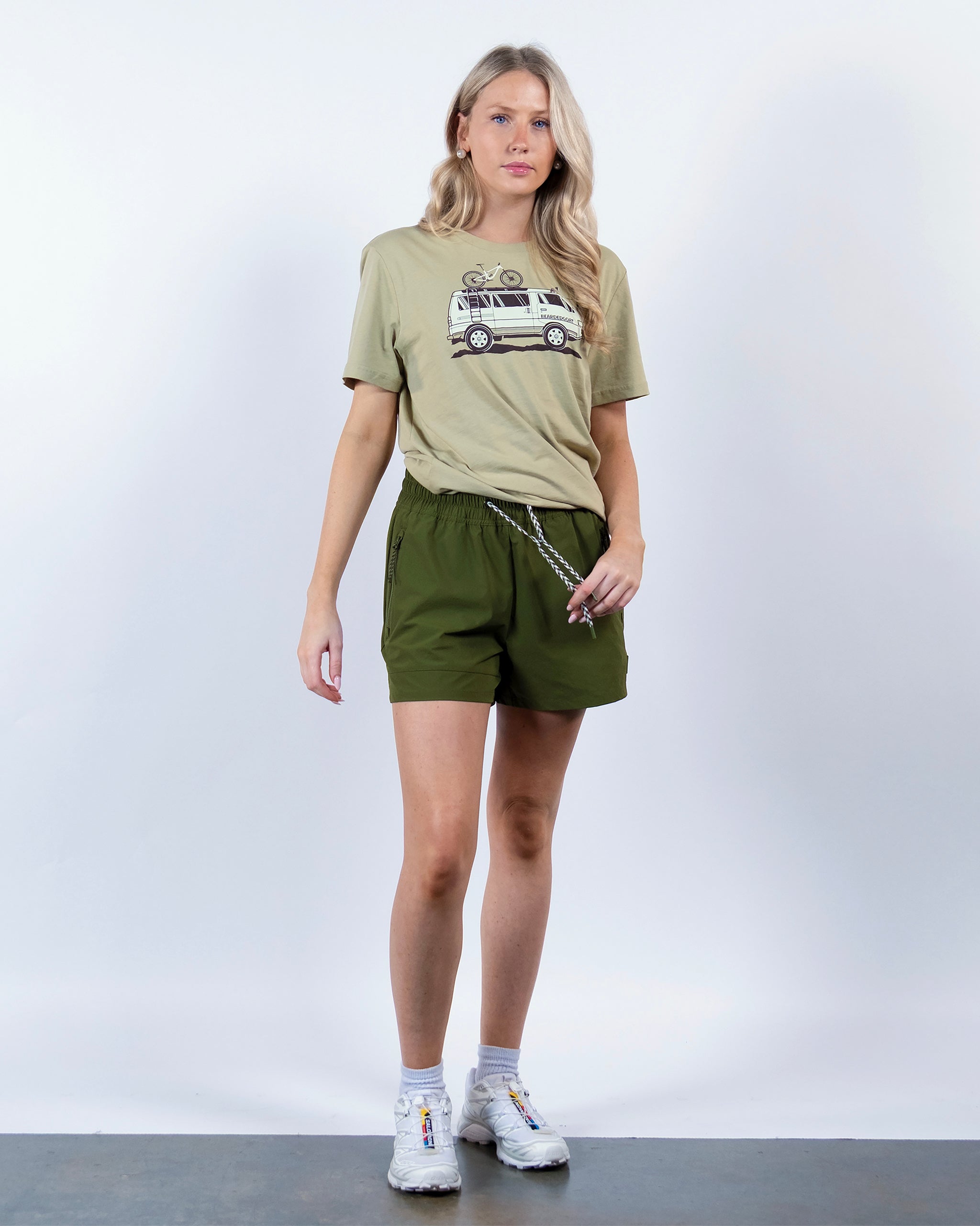 A woman with long blond hair and a neutral expression stands against a white background wearing BEARDEDGOAT Women's Alpina Short in green, a beige van graphic T-shirt, white socks, and white sneakers.
