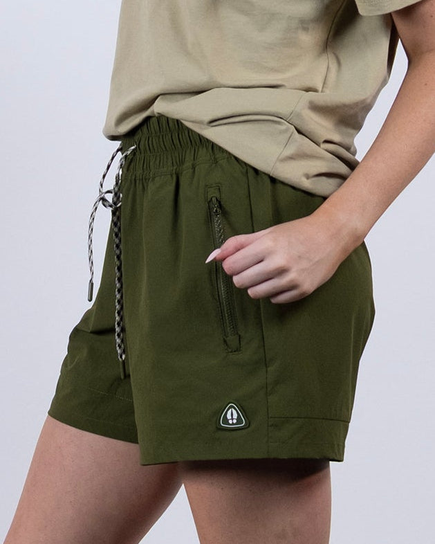 A woman with long blonde hair stands sideways against a plain background, wearing a beige graphic tee and BEARDEDGOAT Women's Alpina Short made from ripstop fabric, paired with white socks and sneakers.