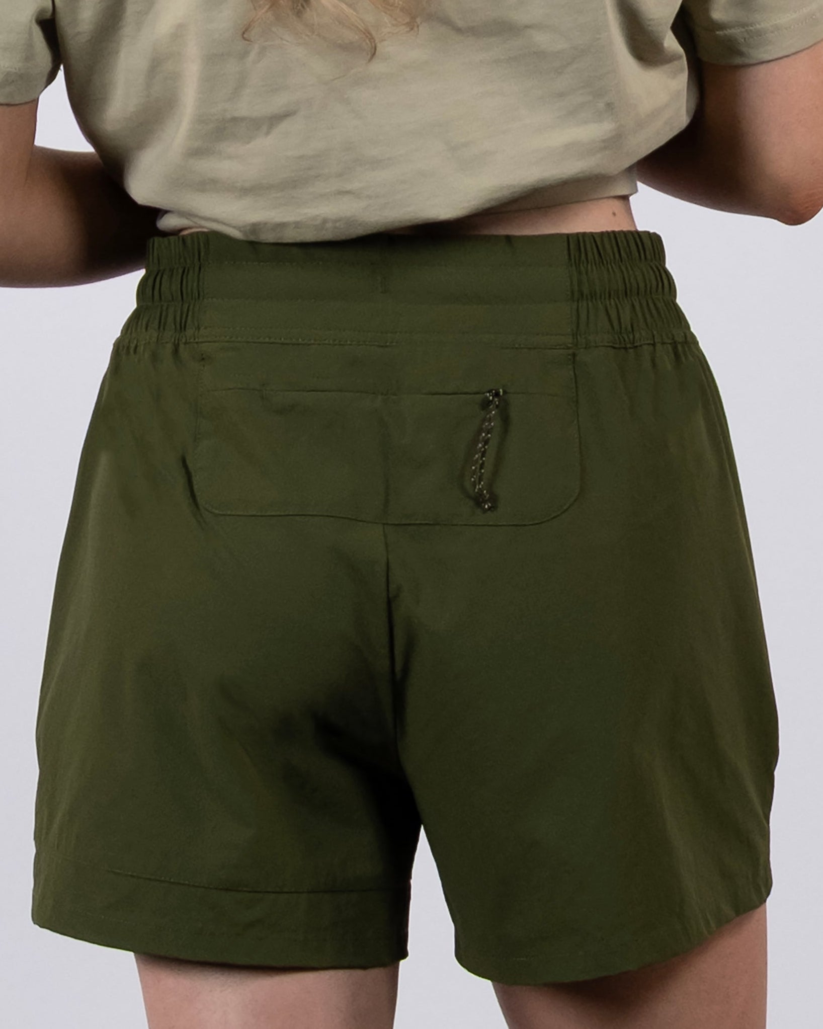 A woman with long, wavy blonde hair is seen from behind wearing a short-sleeve beige shirt and BEARDEDGOAT Women's Alpina Short, which is high-waisted, made of ripstop fabric, and features an elastic waistband and zippered pockets.
