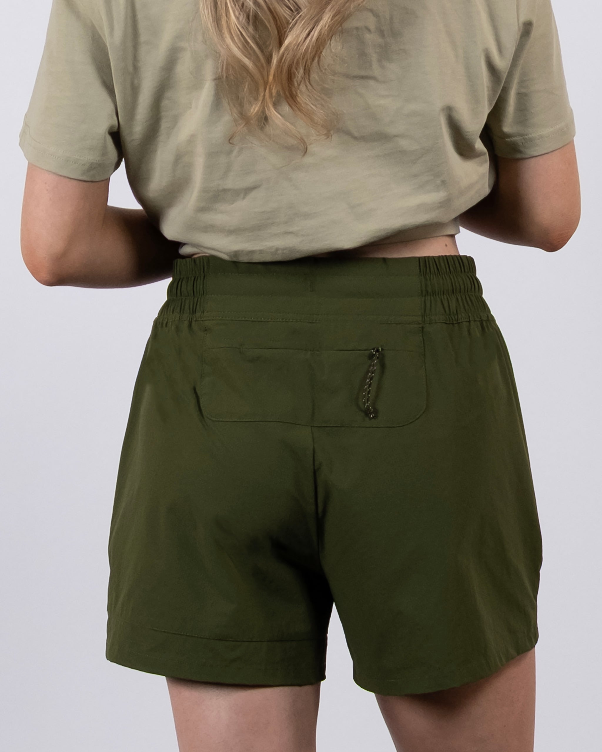 A woman with long, wavy blonde hair is seen from behind wearing a short-sleeve beige shirt and BEARDEDGOAT Women's Alpina Short, which is high-waisted, made of ripstop fabric, and features an elastic waistband and zippered pockets.