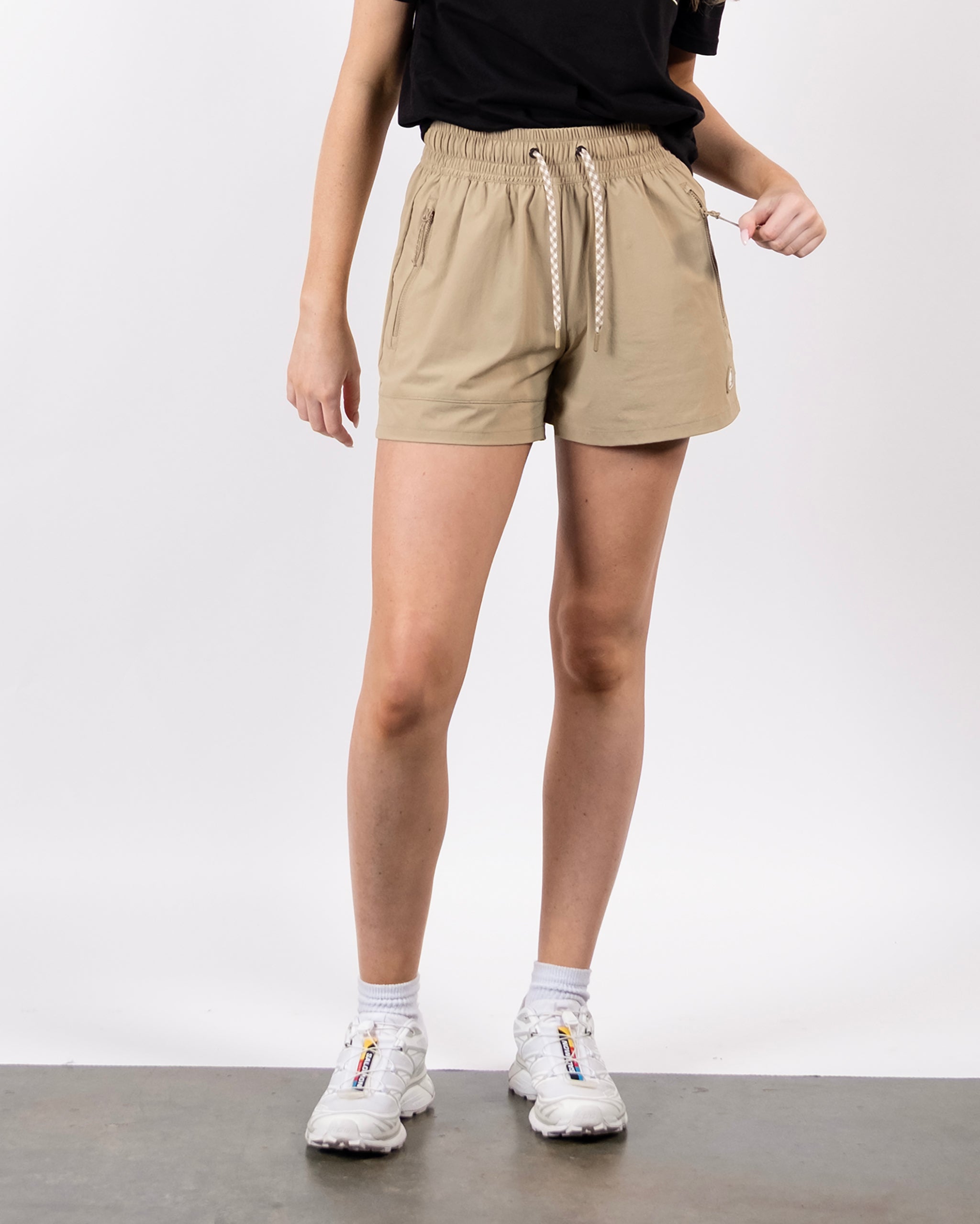 A person stands against a plain white background wearing a black t-shirt, BEARDEDGOAT Women's Alpina Short with zippered pockets, white socks, and white sneakers. Their face is not visible.