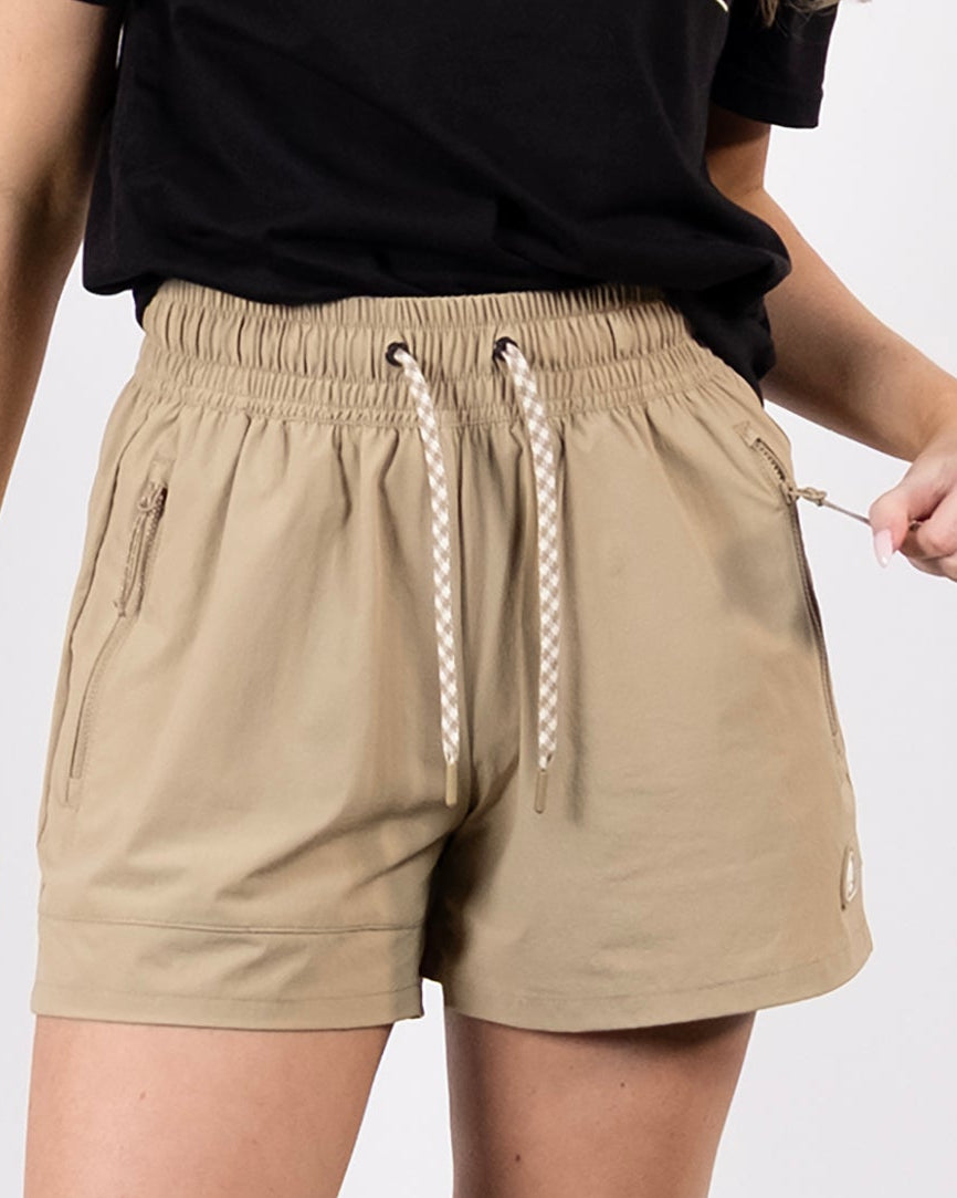 A person stands against a plain white background wearing a black t-shirt, BEARDEDGOAT Women's Alpina Short with zippered pockets, white socks, and white sneakers. Their face is not visible.