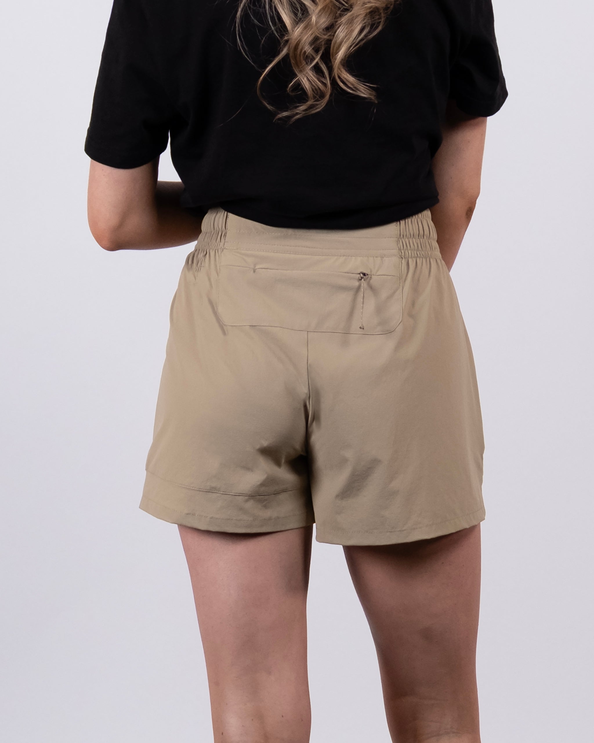 A person with long, wavy blonde hair is seen from the back, wearing a black shirt and BEARDEDGOAT Women's Alpina Short—beige, high-waisted adventure shorts with an elastic waistband and small back pocket—against a plain light background.