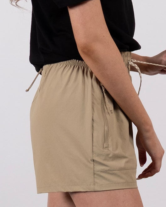 A woman with long blonde hair stands sideways against a plain background, wearing a black t-shirt and BEARDEDGOAT Women's Alpina Short—beige, high-waisted adventure shorts with zippered pockets—paired with white socks and sneakers.