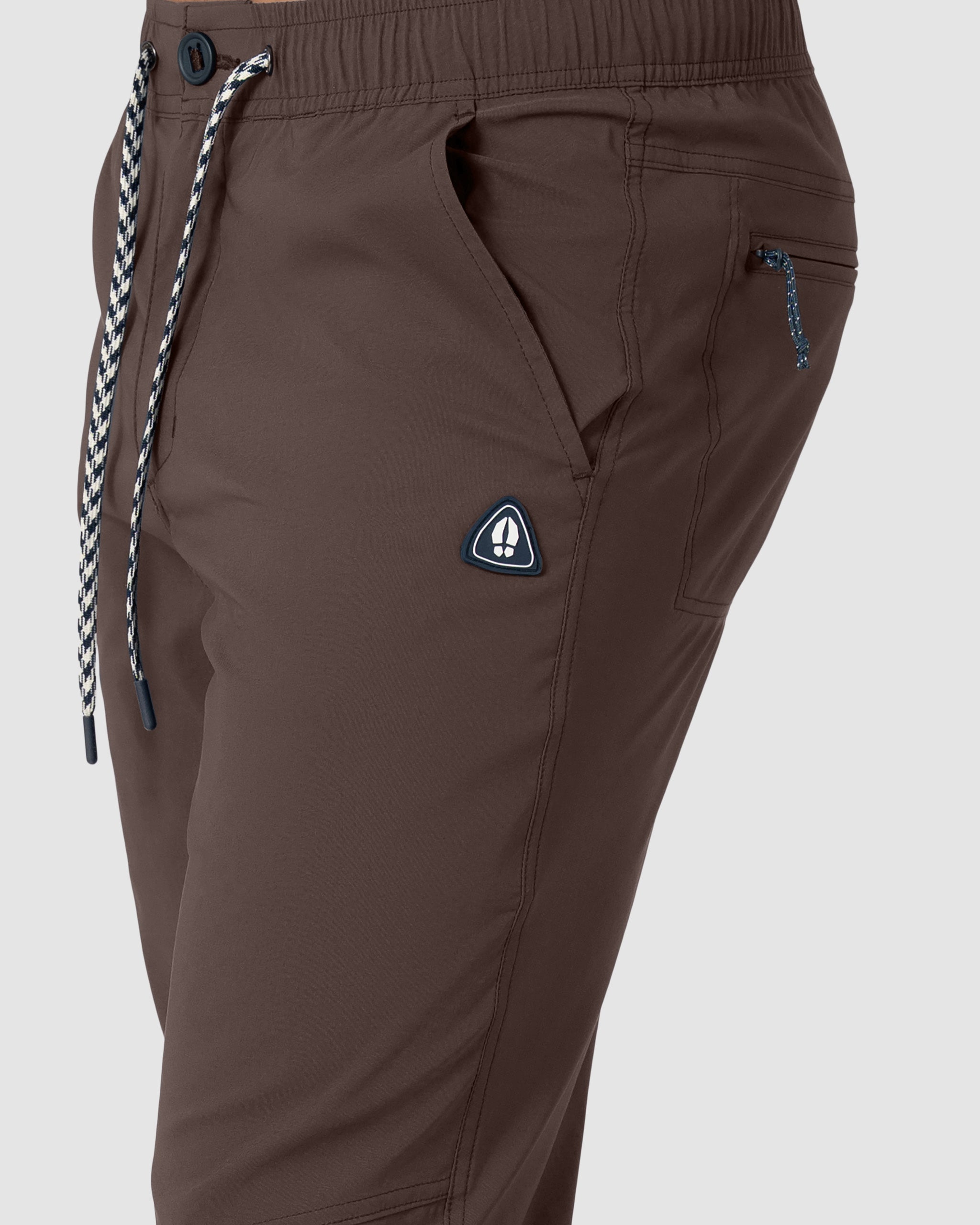 Close-up of a person wearing BEARDEDGOAT's NEW-—ROVER™ Pant in brown, featuring a black-and-white drawstring, zippered pocket, and a small triangular logo patch near the pocket.