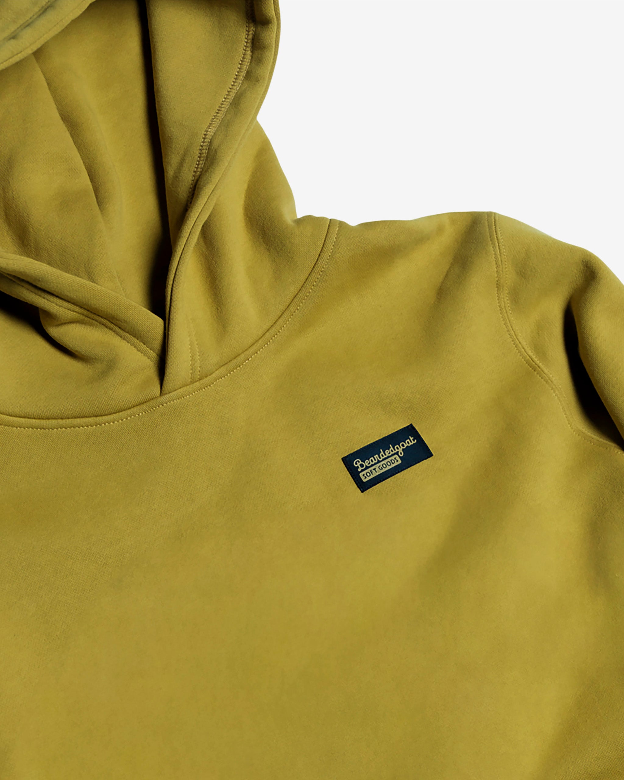 A close-up of the beardedgoat Women's Overnight™ Hoodie in mustard yellow, made from premium cotton fleece. It has a relaxed fit and a small navy chest patch with "ParadeSport" in white, shown laid flat on a white background.