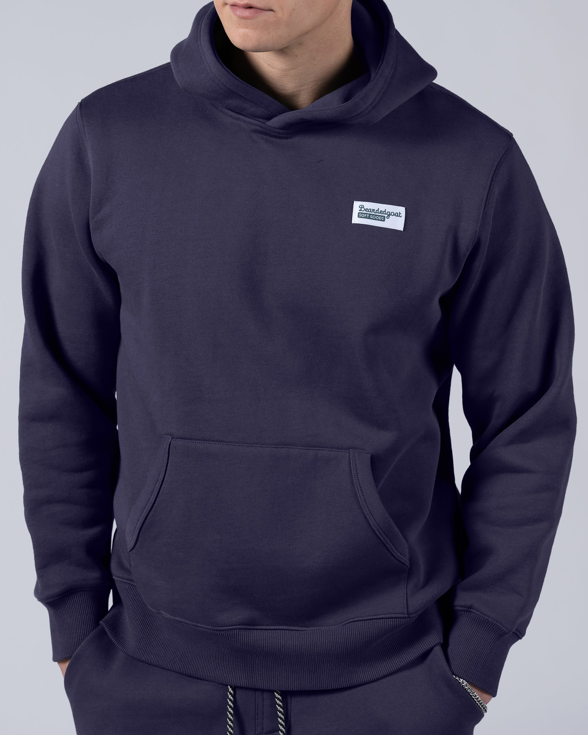 A person wears the beardedgoat Men's Void Hoodie in dark blue heavyweight cotton fleece with a small white text patch on the chest, hood up and hands in the front pocket, set against a plain light gray background.