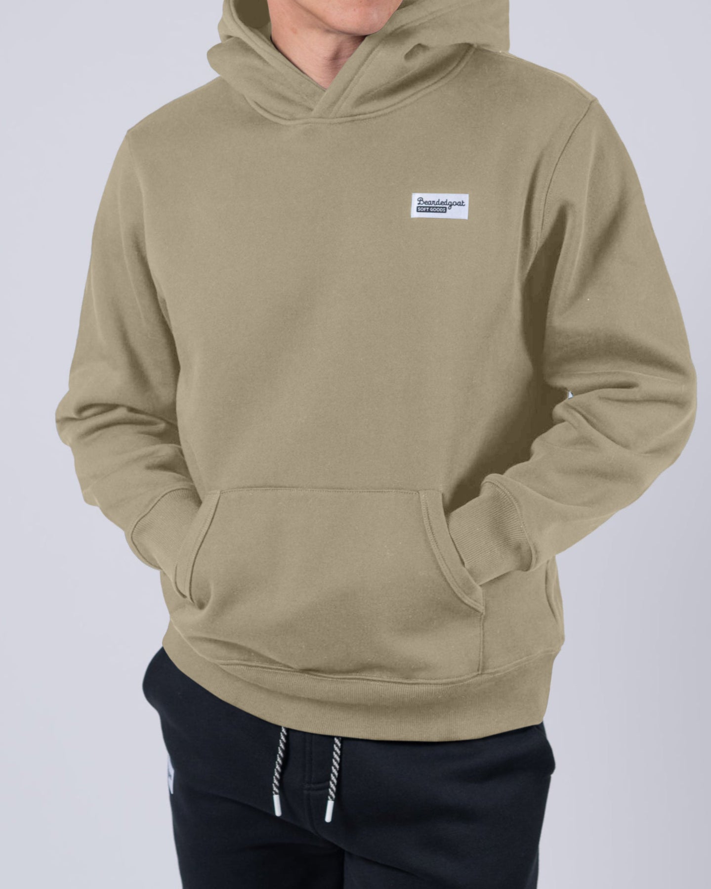 A man stands against a plain background wearing the beardedgoat Men's Void Hoodie in light brown with a small chest logo and black drawstring pants, hands in the hoodie pocket.