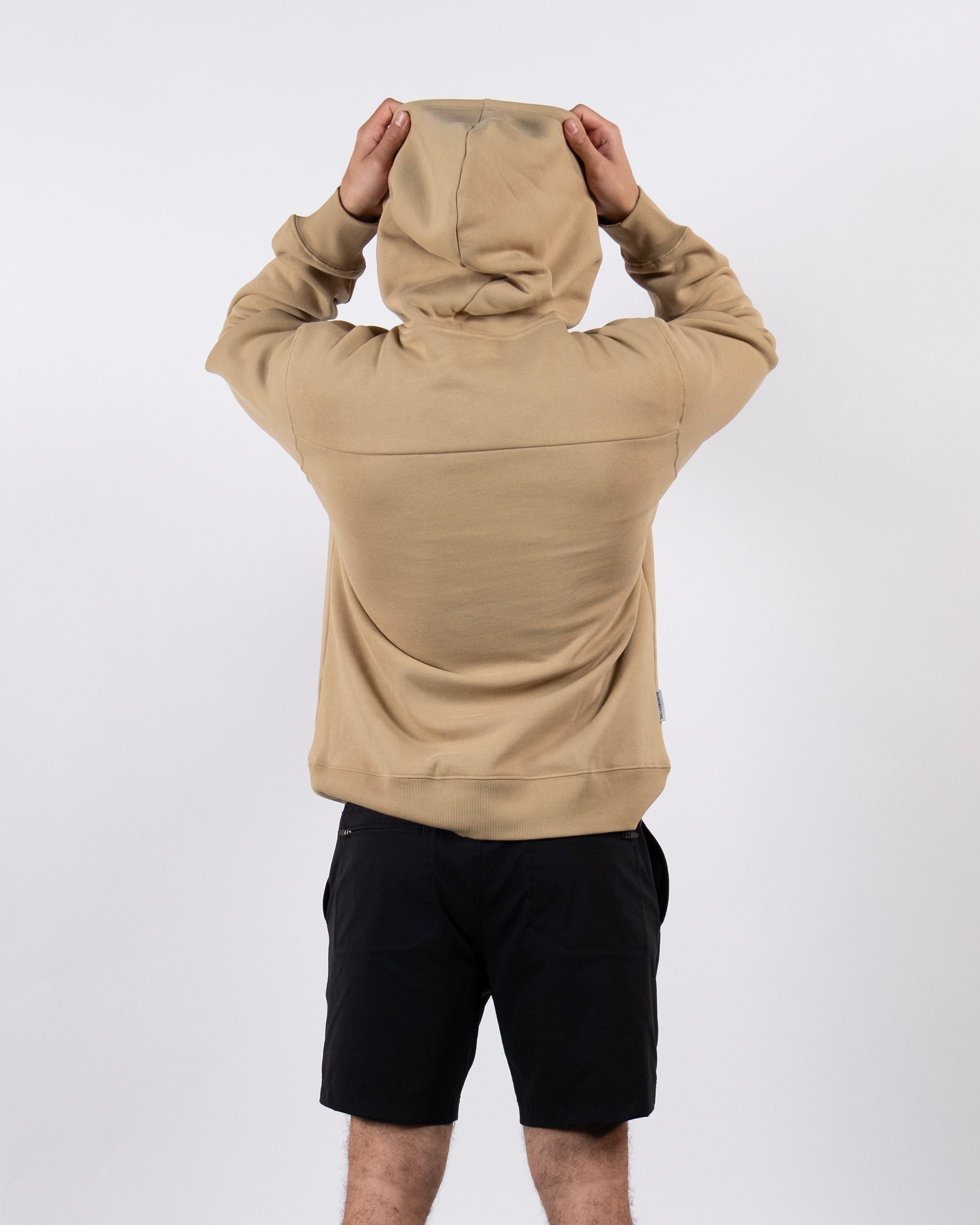 A person in a tan beardedgoat The VOID™ Hoodie, made from heavyweight cotton fleece, stands with the hood up and black shorts, facing away from the camera against a plain white background.